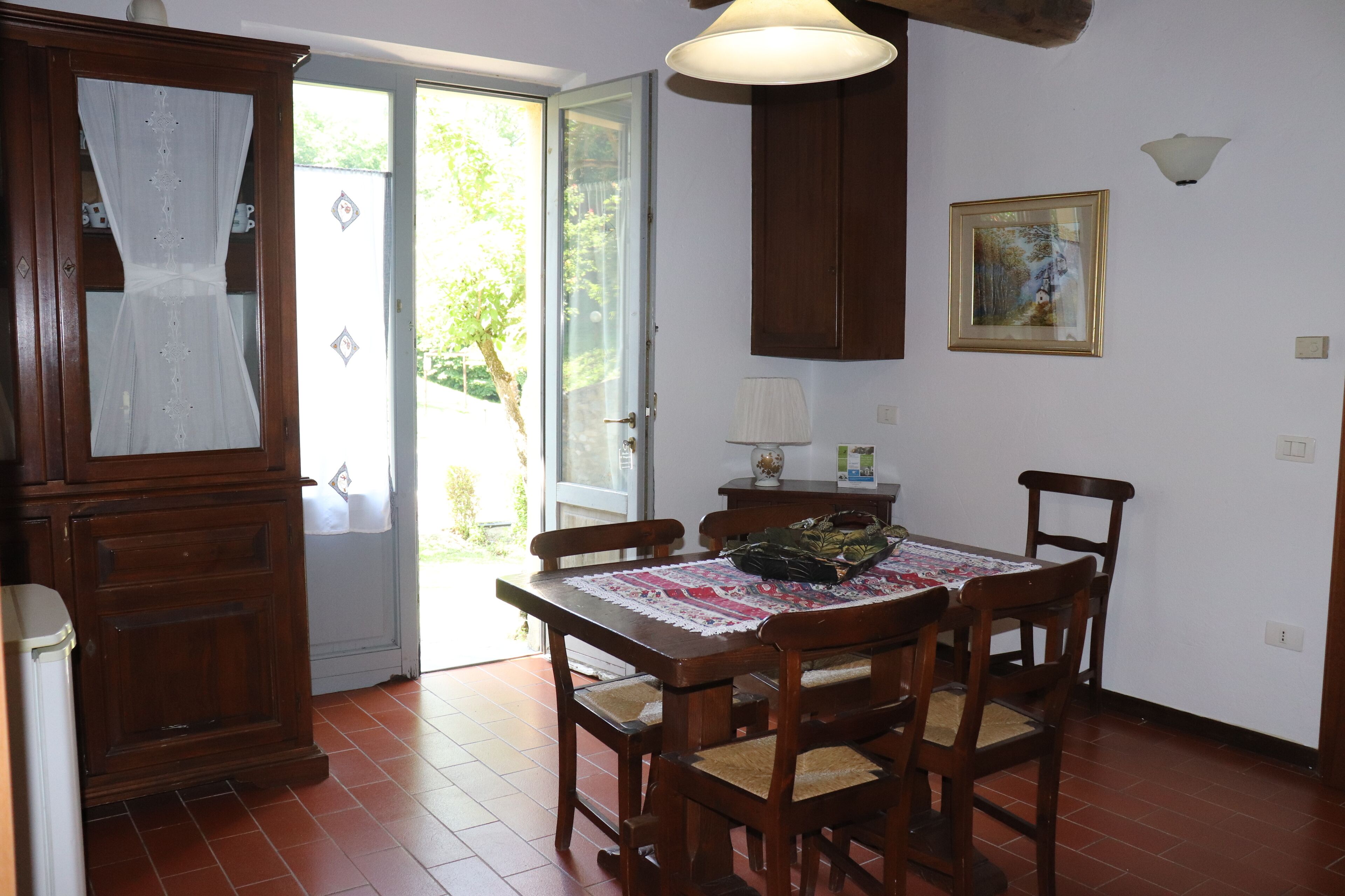 A dining area with a wooden table, chairs, and a lace tablecloth. There is a cabinet with a mirror, a door leading outside, and a wall-mounted lamp.