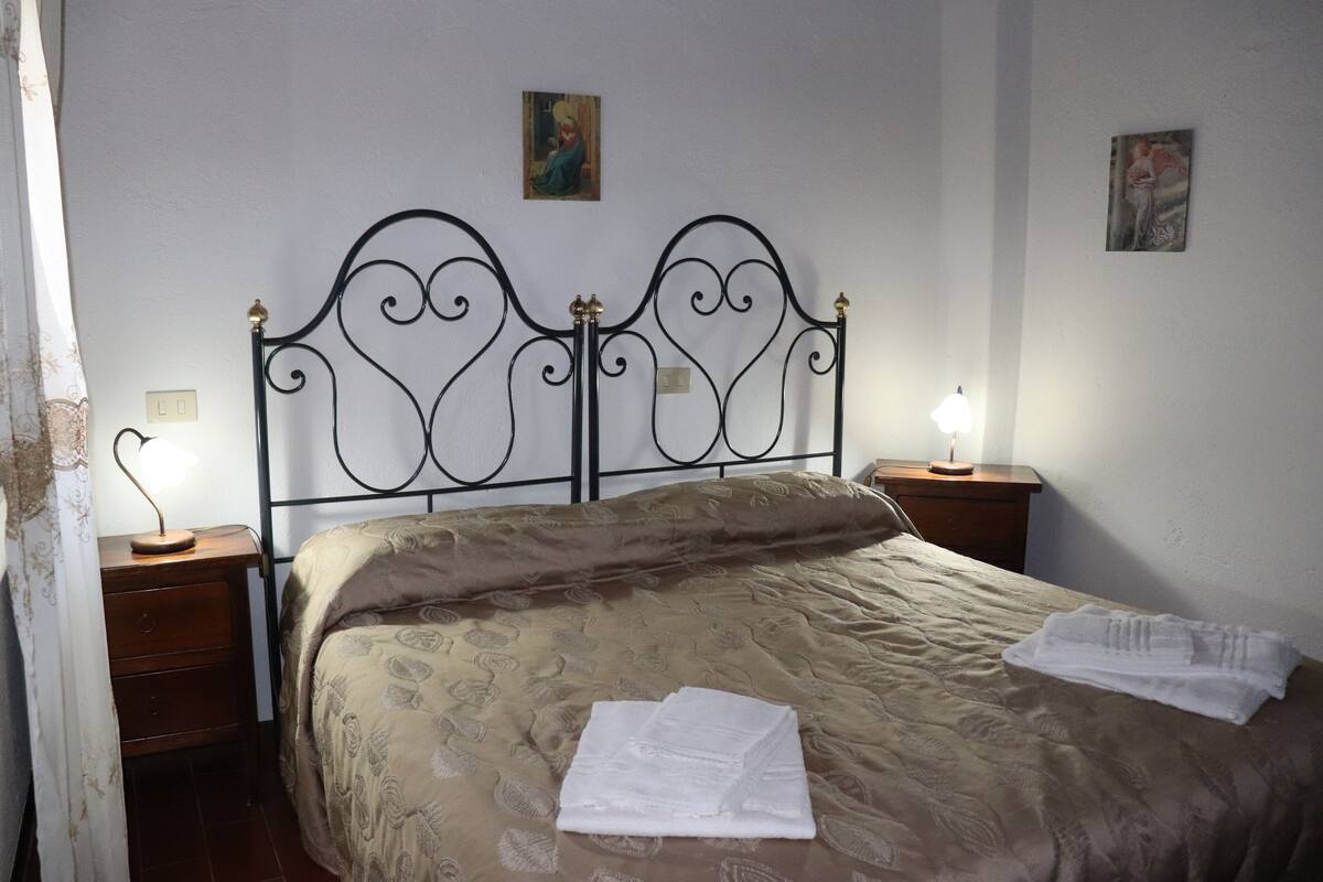 A neatly made bed with a patterned bedspread and a folded towel on the side. There are two bedside tables with lamps, and artwork is visible on the walls.
