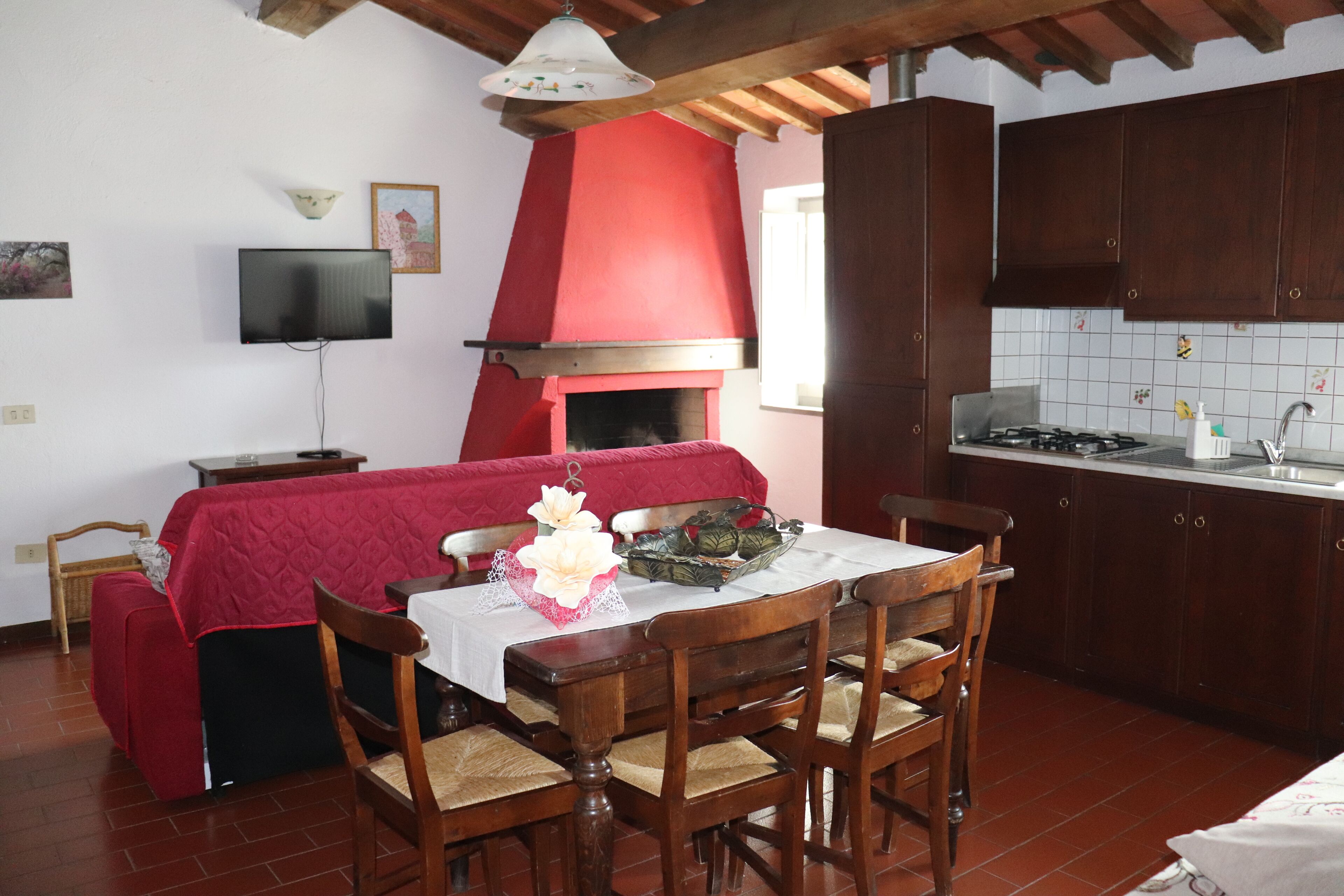A dining area with a red-covered bench, a table with chairs, a TV mounted on the wall, and a kitchen with dark wooden cabinets and a tiled backsplash.