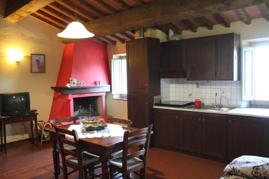 Een keuken met houten kastjes, een rode schoorsteen, een eettafel met stoelen en een televisie op een bijzettafeltje.