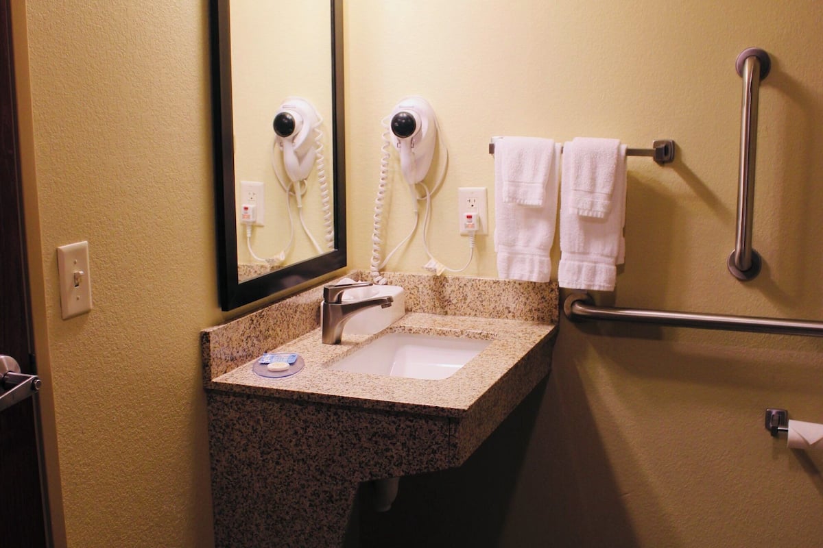 Un baño con lavamanos, espejo, secador de pelo y secador de manos. Hay un cubo de basura y un perchero con toallas.