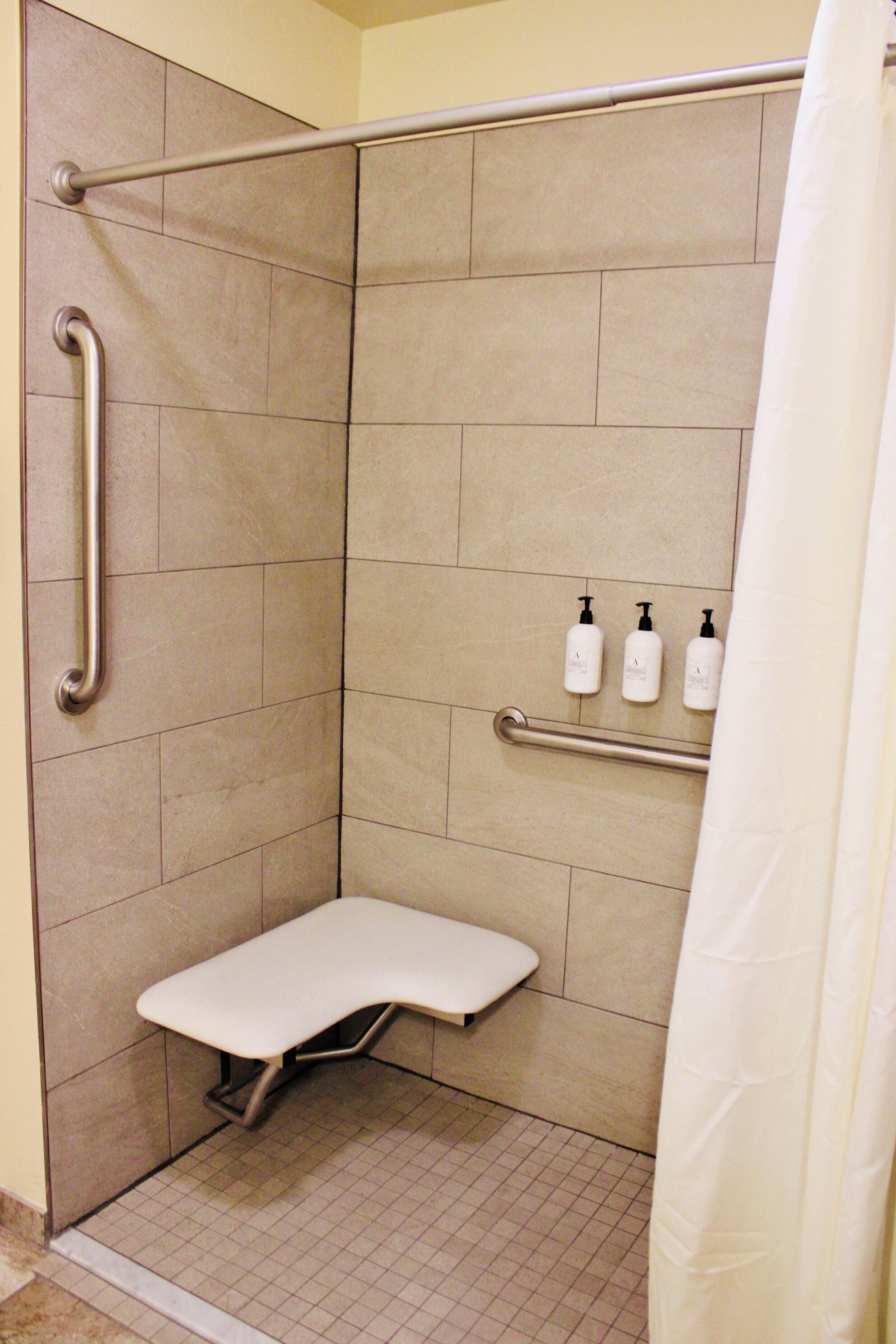 A shower stall with a grab bar, a bench, and three bottles of shampoo on the wall.
