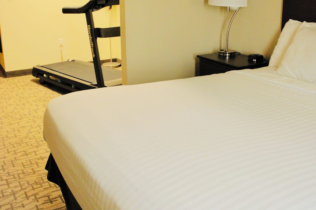 Habitación de hotel con una cama grande, un escritorio con lámpara, una caminadora y un cuadro en la pared.