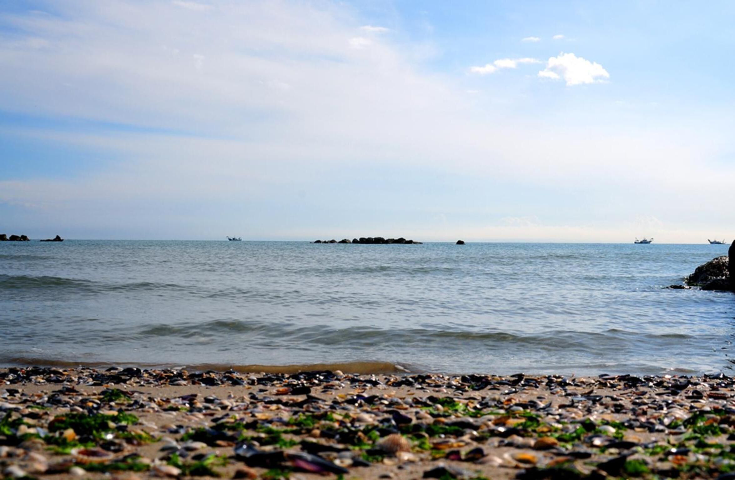 Een strand met kiezelstenen en rotsen, kalm water en verre boten.