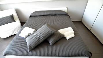 A neatly made bed with grey bedding and pillows, a white bedside table, and a white chair.