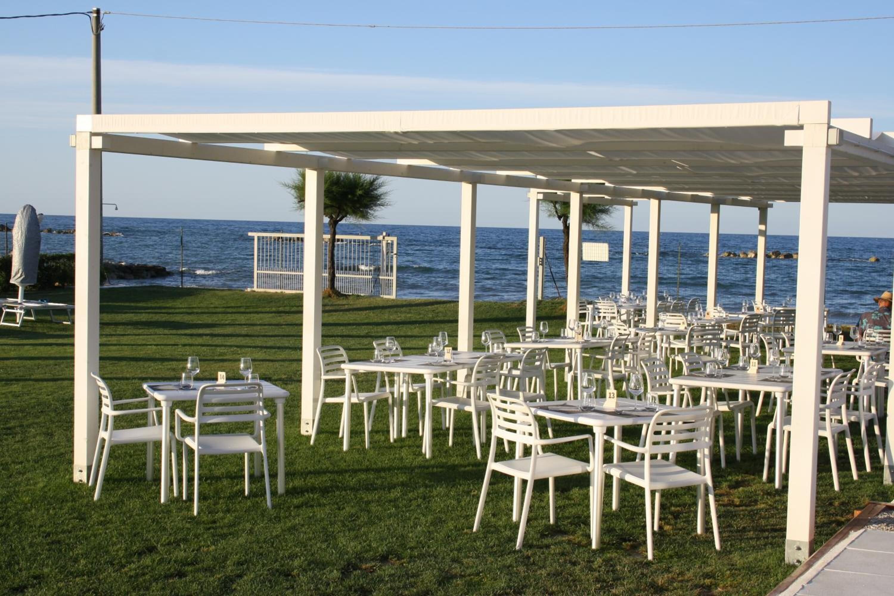 Un área de comedor al aire libre con mesas y sillas blancas, una pérgola cubierta y vistas al mar.