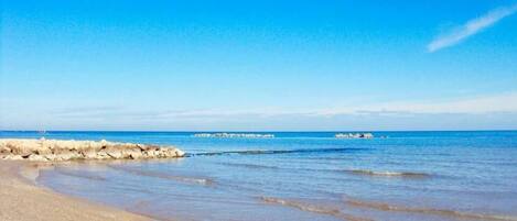 A beach with clear blue water, a rocky breakwater, and a distant ship.