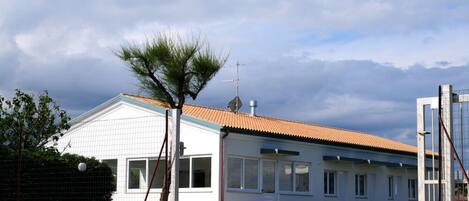 Un bâtiment d’un seul étage, avec un toit recouvert de tuiles, une antenne parabolique et un petit arbre devant.