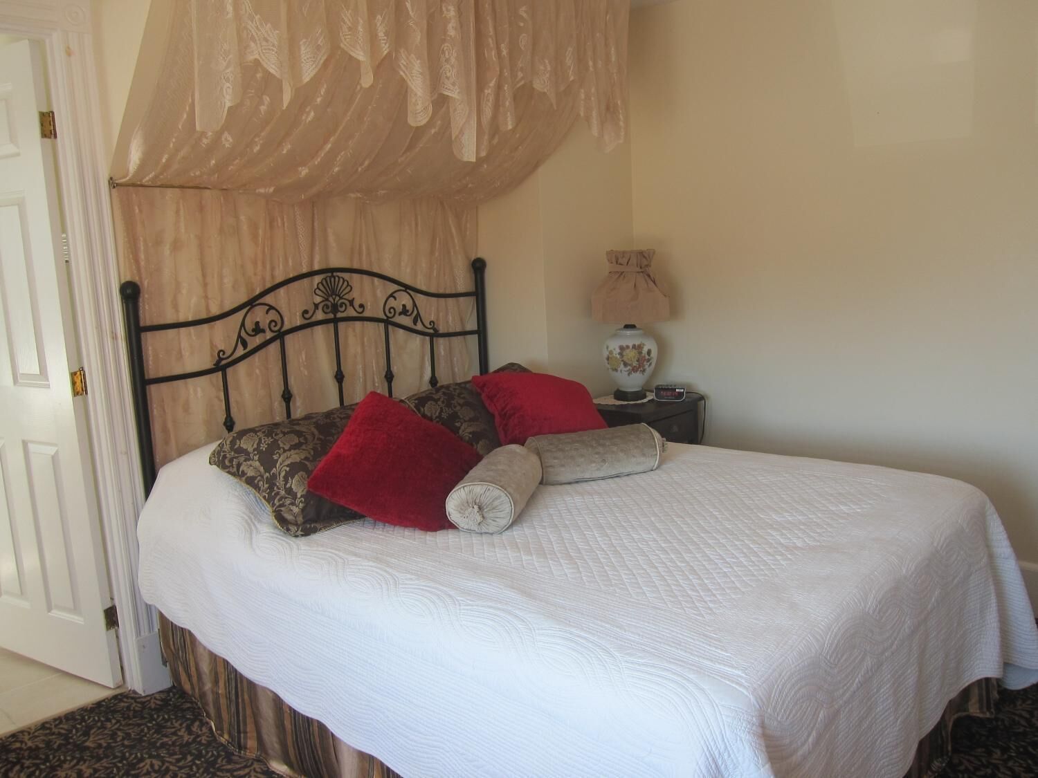 A neatly made bed with a white bedspread and two red pillows, a black metal headboard, and a bedside table with a lamp.
