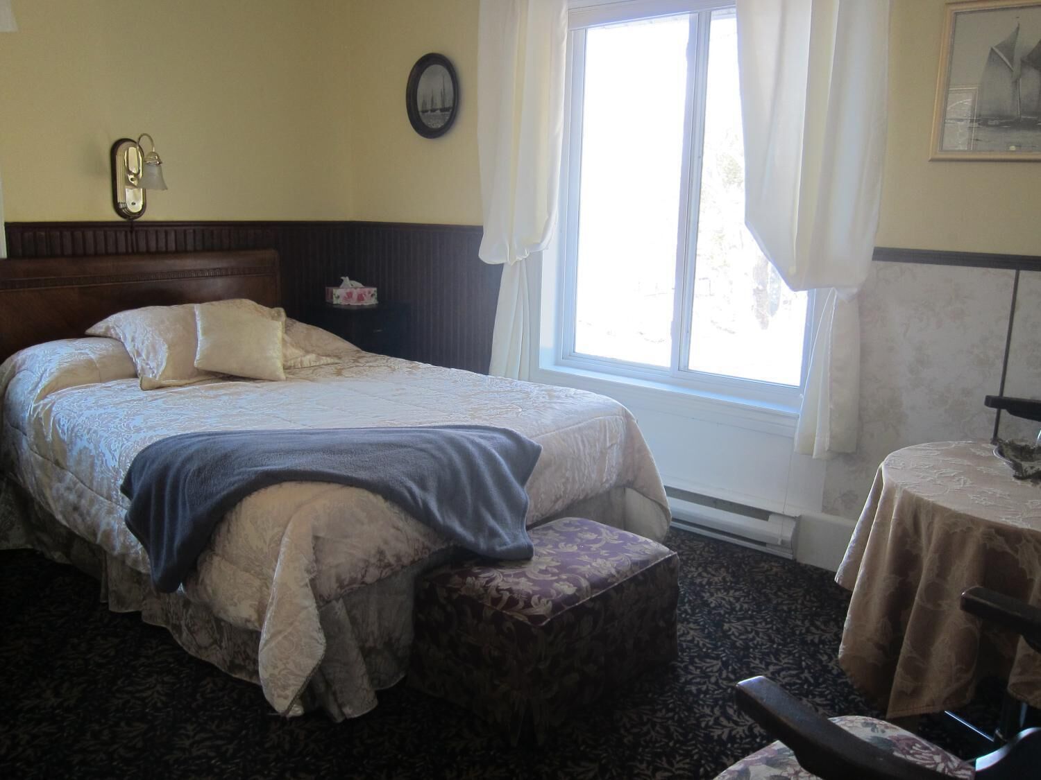Un lit bien fait, avec une tête de lit, une table de chevet avec une lampe, une fenêtre avec des rideaux et une petite table ronde accompagnée d’une chaise.
