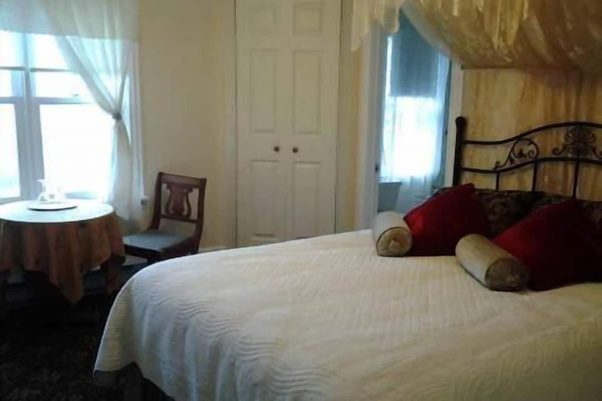 A neatly made bed with red and beige pillows, a small round table with a chair, and a door in the background.