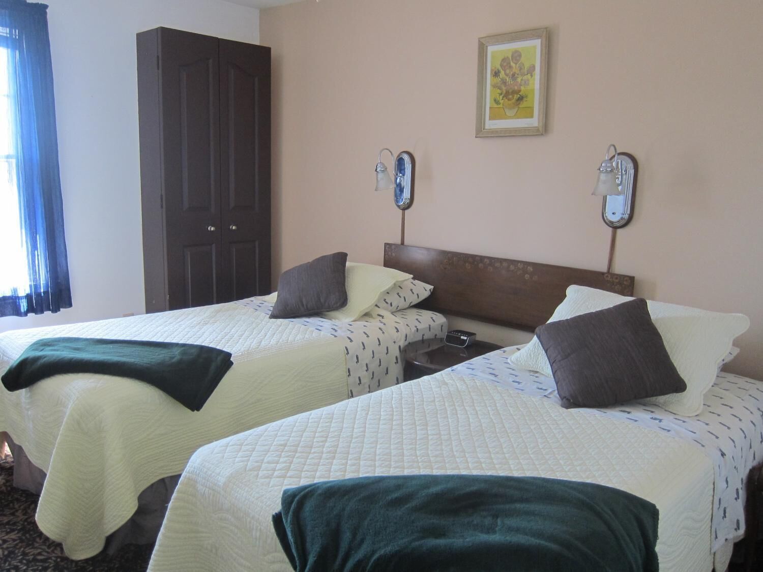Habitación de hotel con dos camas, un armario, una mesita de noche con lámpara y un cuadro en la pared.