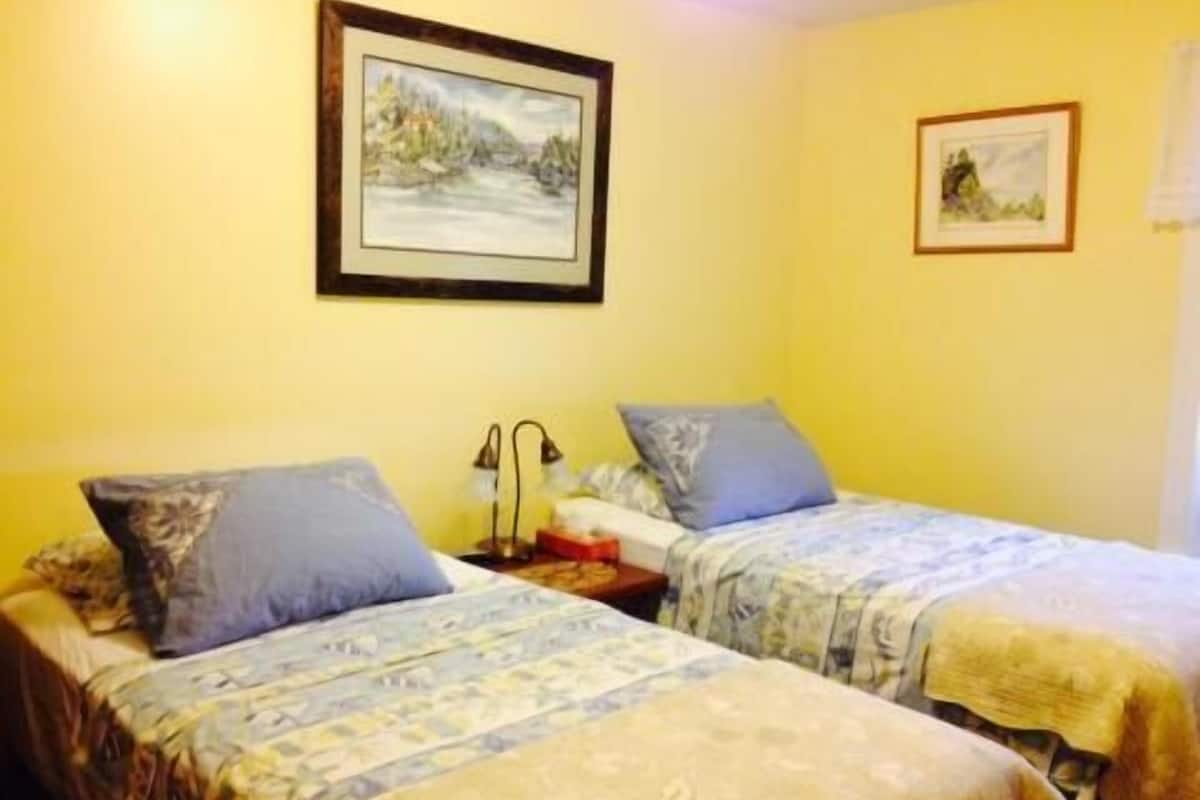 A room with two beds, each with a patterned bedspread and pillows, a nightstand with a lamp, and two framed pictures on the wall.