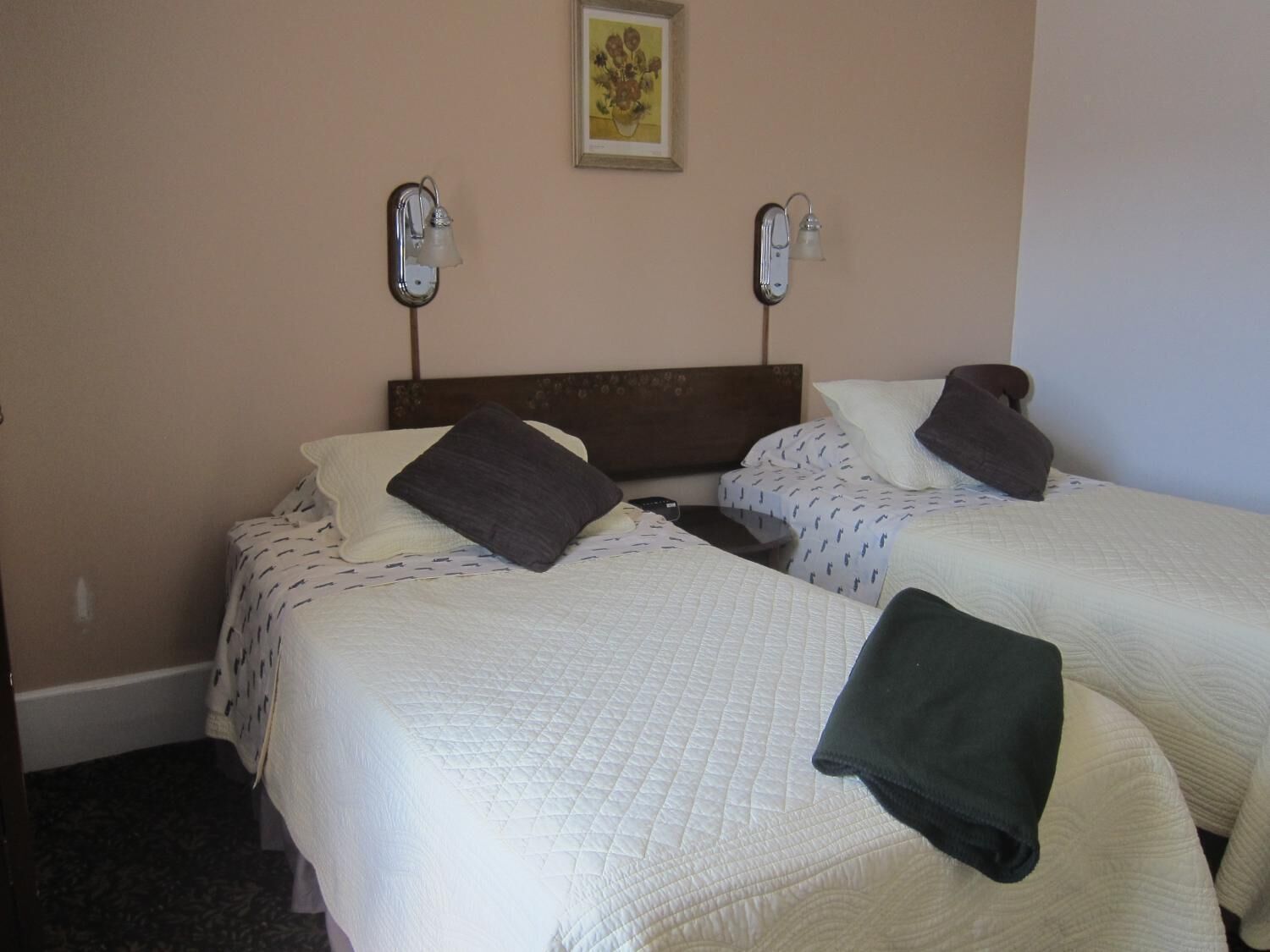 A hotel room with two beds, each with a white quilted bedspread and dark pillows. There are two wall-mounted lamps on either side of the bed.