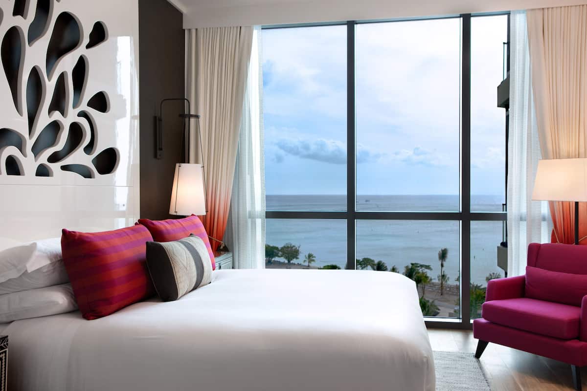 A hotel room with a large bed, a view of the ocean, a red armchair, and a decorative wall panel.