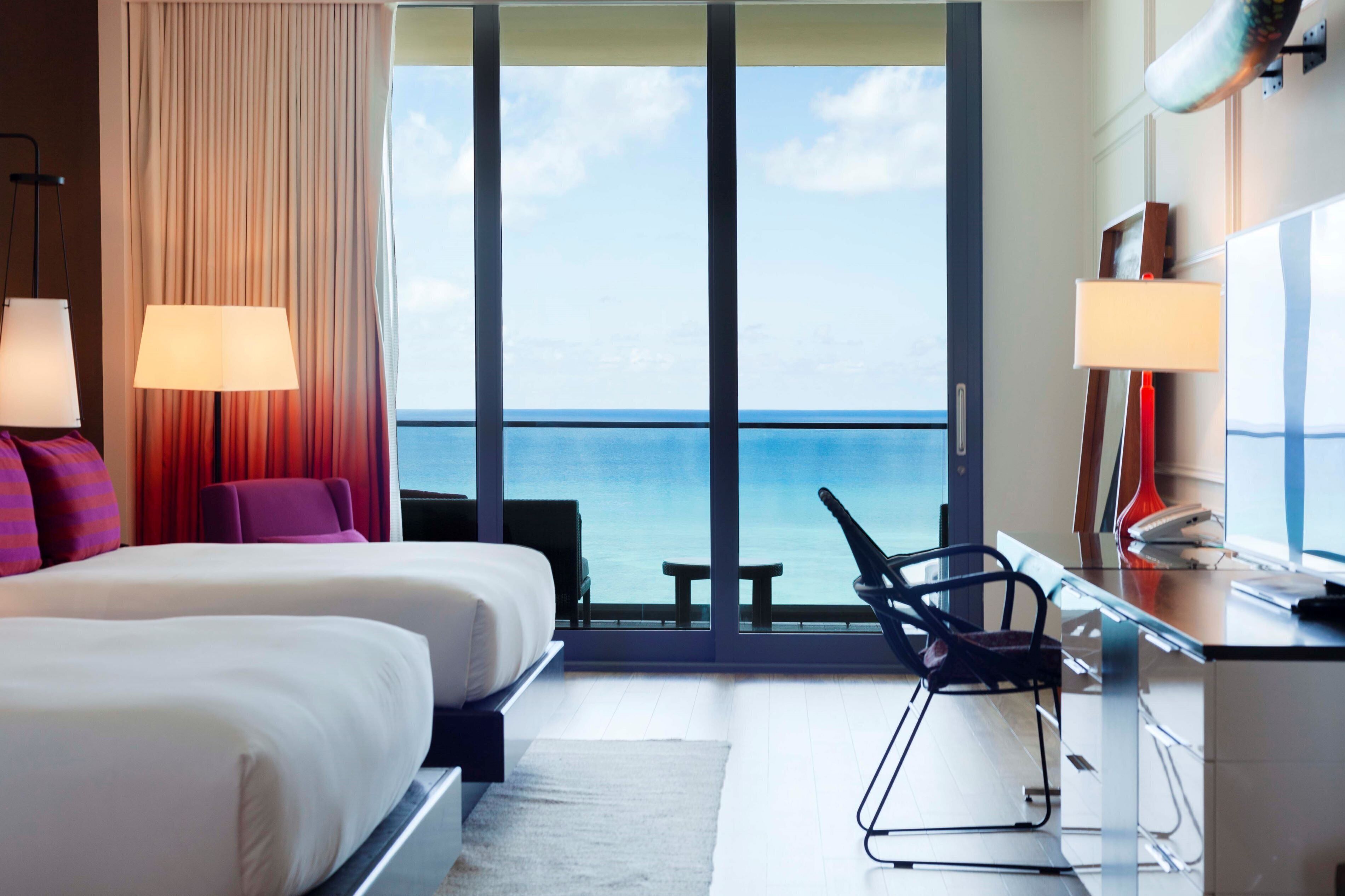 Ein Hotelzimmer mit einem großen Bett, zwei Nachttischlampen, einem Schreibtisch mit Computer, einem Stuhl und Meerblick durch raumhohe Fenster.
