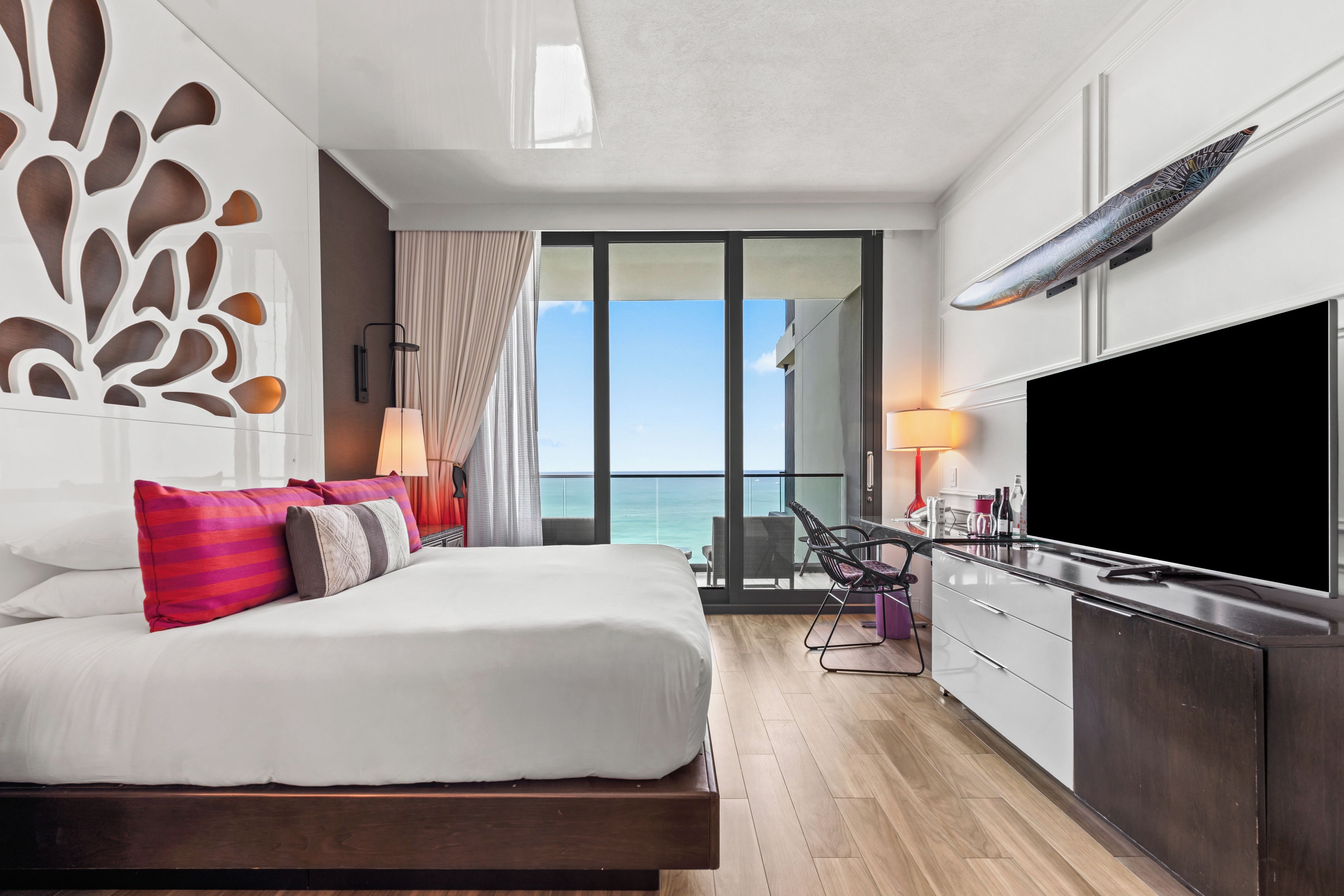 A modern hotel room with a bed, a TV, a balcony with a view of the sea, and a dining area.