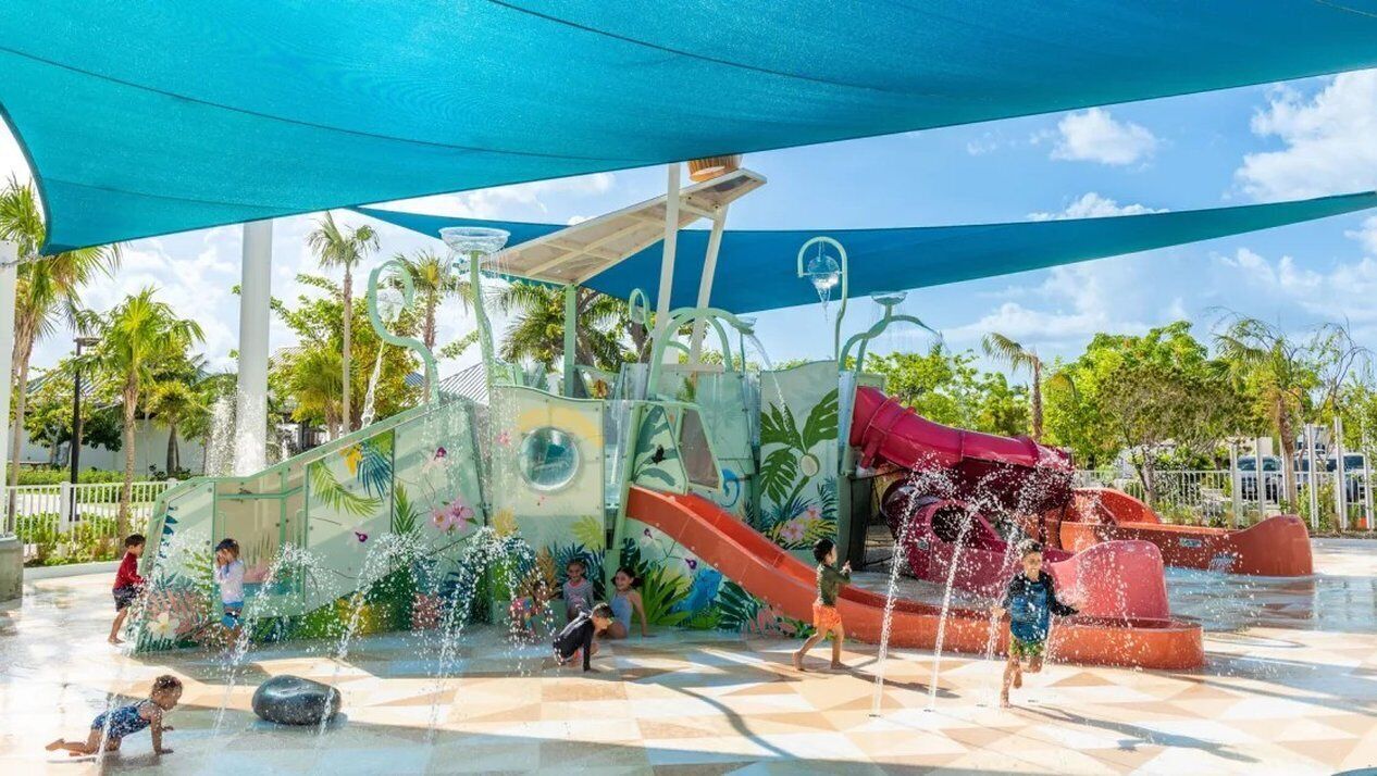 An outdoor water play area with slides and water jets, surrounded by palm trees and a clear sky.
