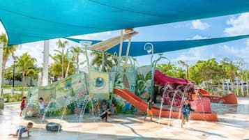 An outdoor water play area with slides and water jets, surrounded by palm trees and a clear sky.