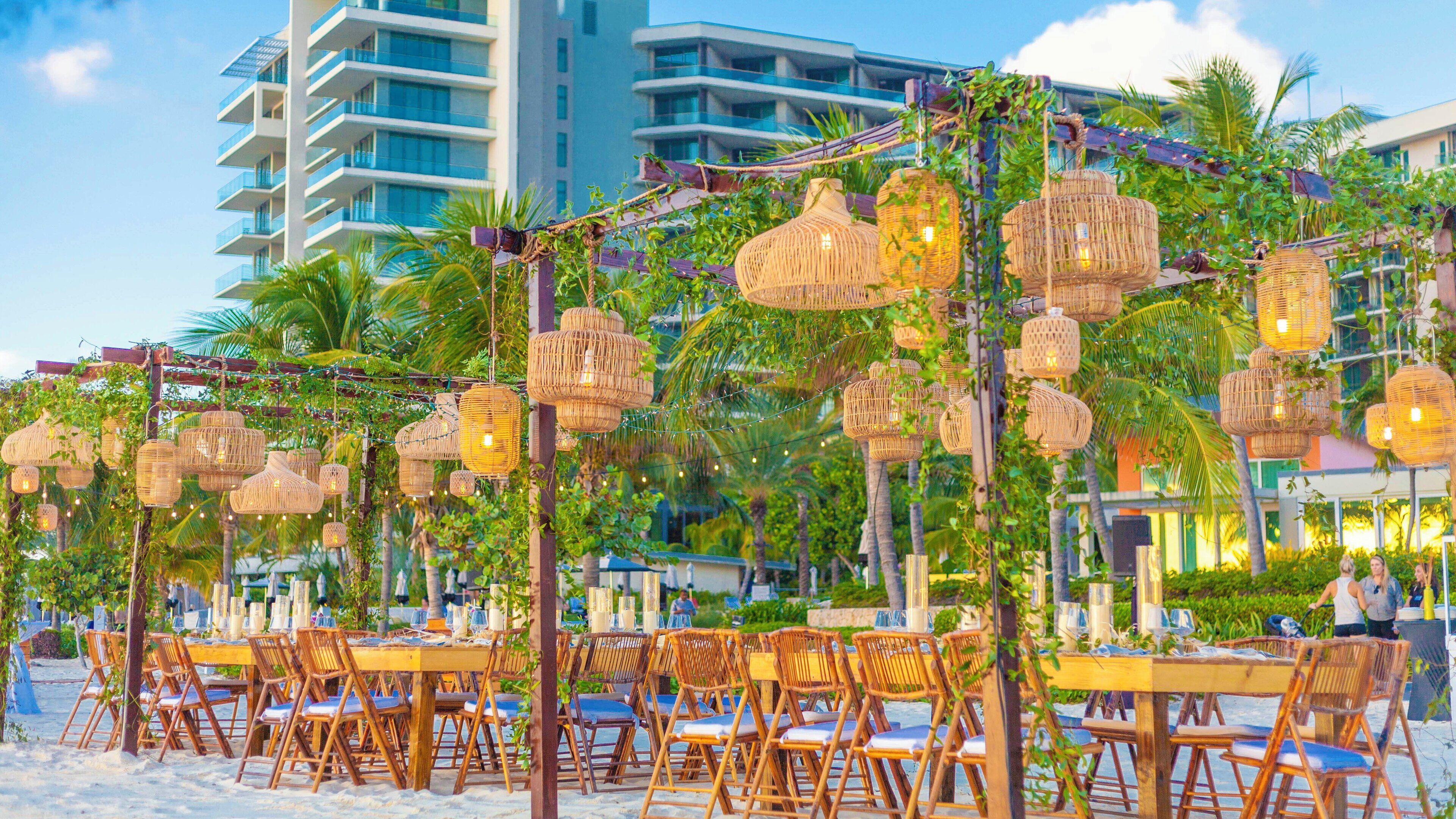 Outdoor dining with wicker lamps and tables, surrounded by greenery and a building in the background.