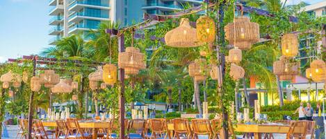 Outdoor dining with wicker lamps and tables, surrounded by greenery and a building in the background.