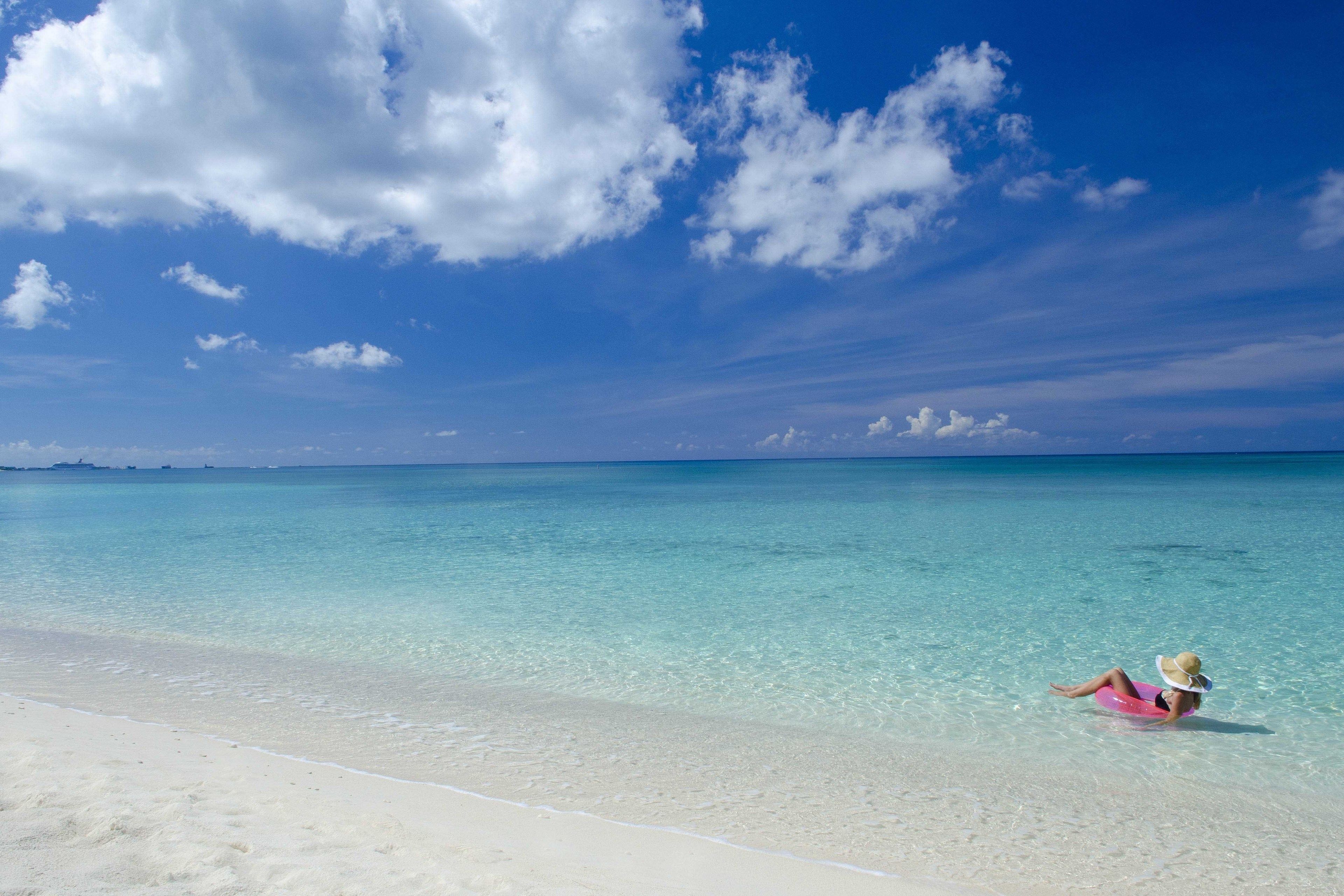 A beach scene with clear turquoise water and a person lying on a pink float.