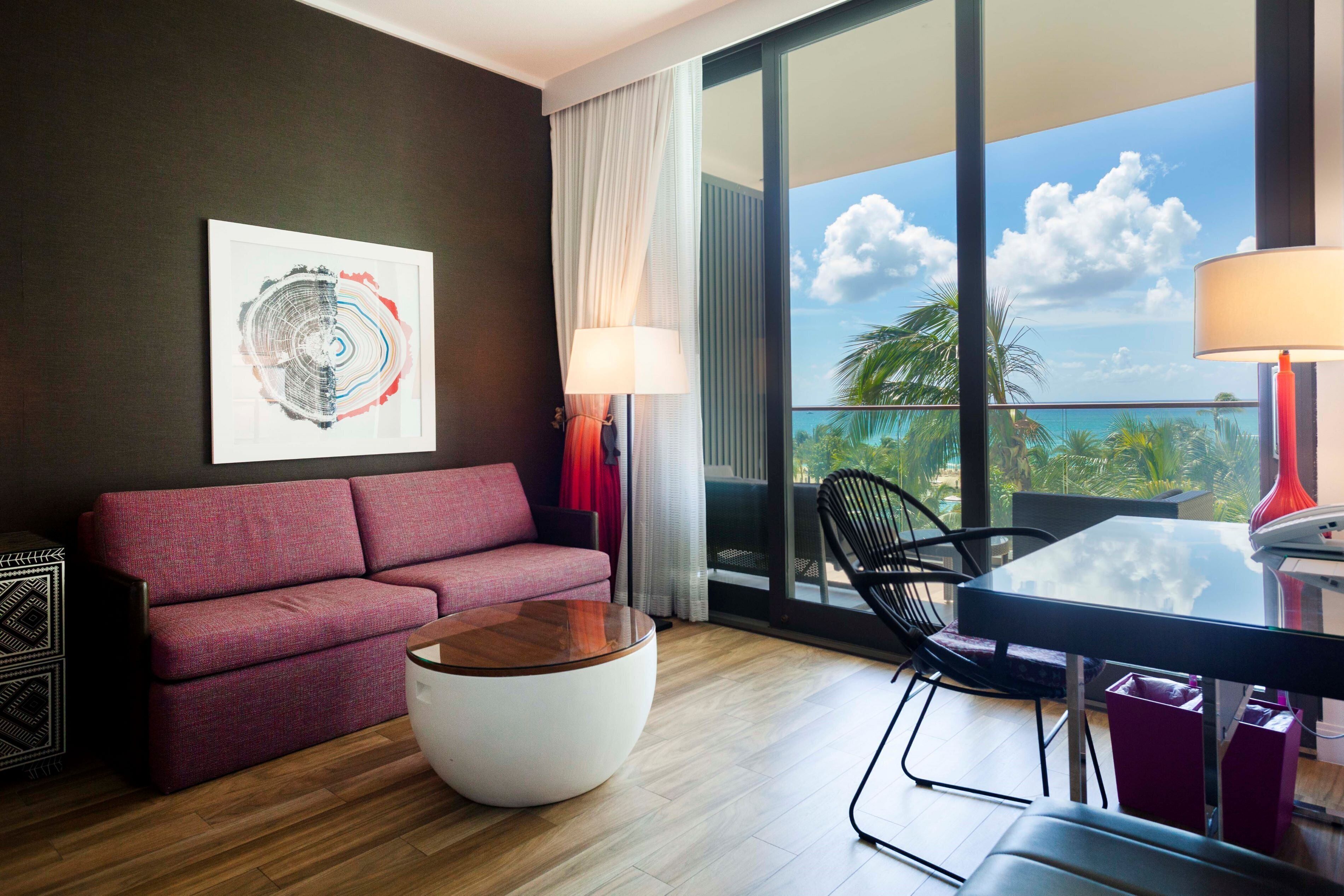Une chambre d’hôtel moderne avec un canapé rouge, une table basse ronde, une table à manger en verre et une grande fenêtre donnant sur des palmiers et le ciel.