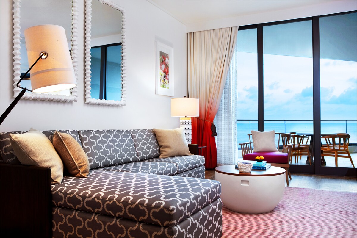 A modern living room with a patterned sofa, a round coffee table, and a large window offering a view of the sea.