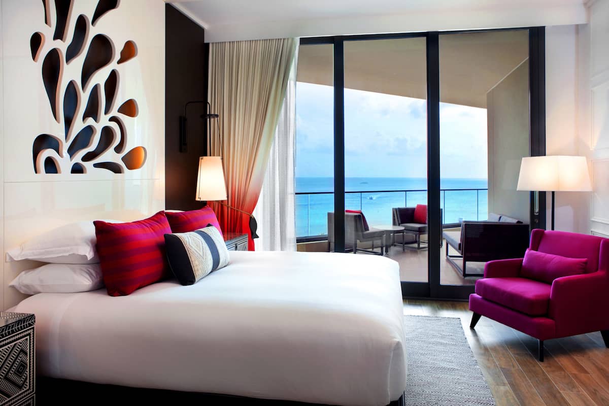 A hotel room with a large bed, a red armchair, and a view of the ocean through a sliding door.