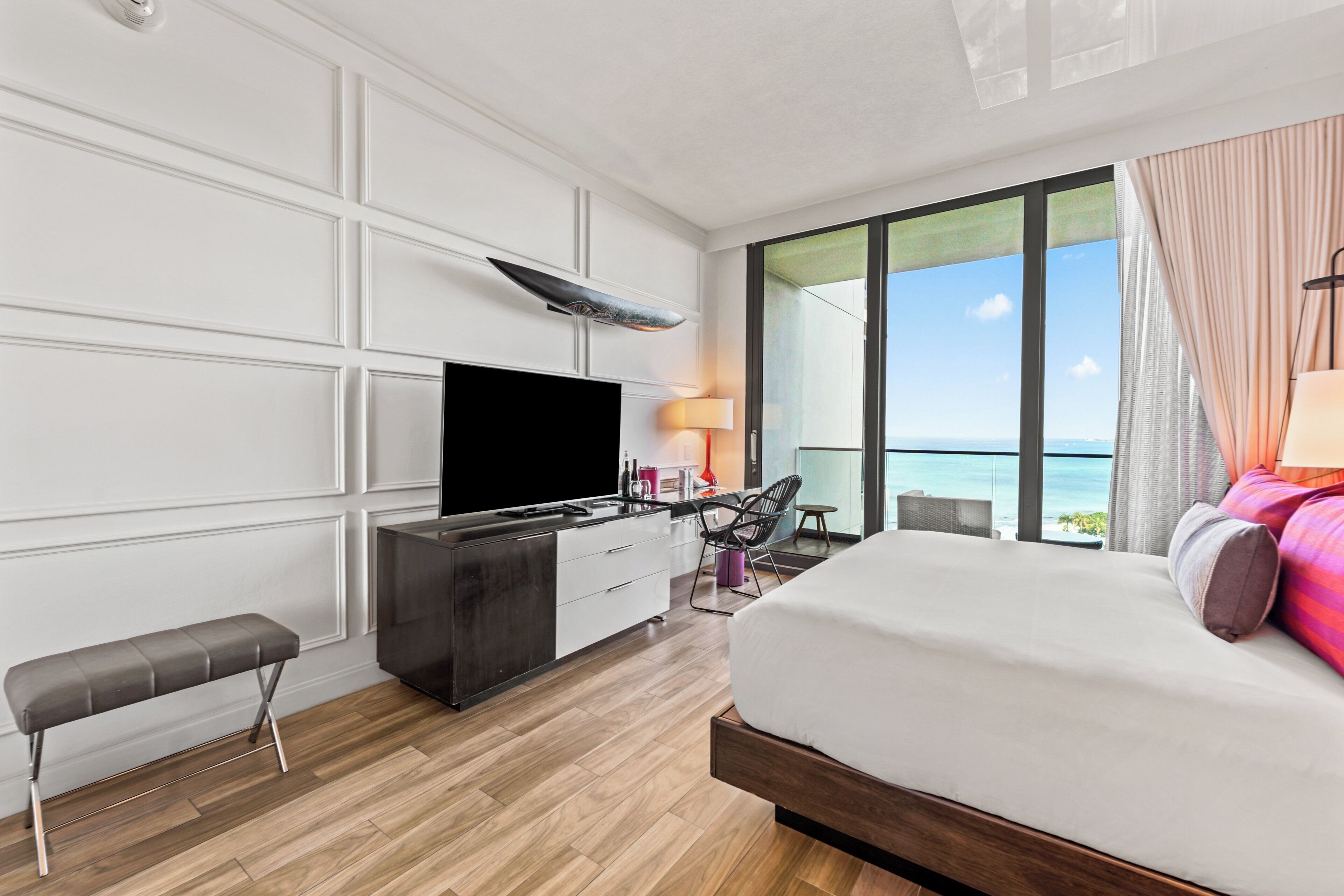 A modern hotel room with a large bed, a flat-screen TV, a desk with a chair, and a view of the ocean.