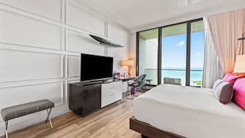 A modern hotel room with a large bed, a flat-screen TV, a desk with a chair, and a view of the ocean.
