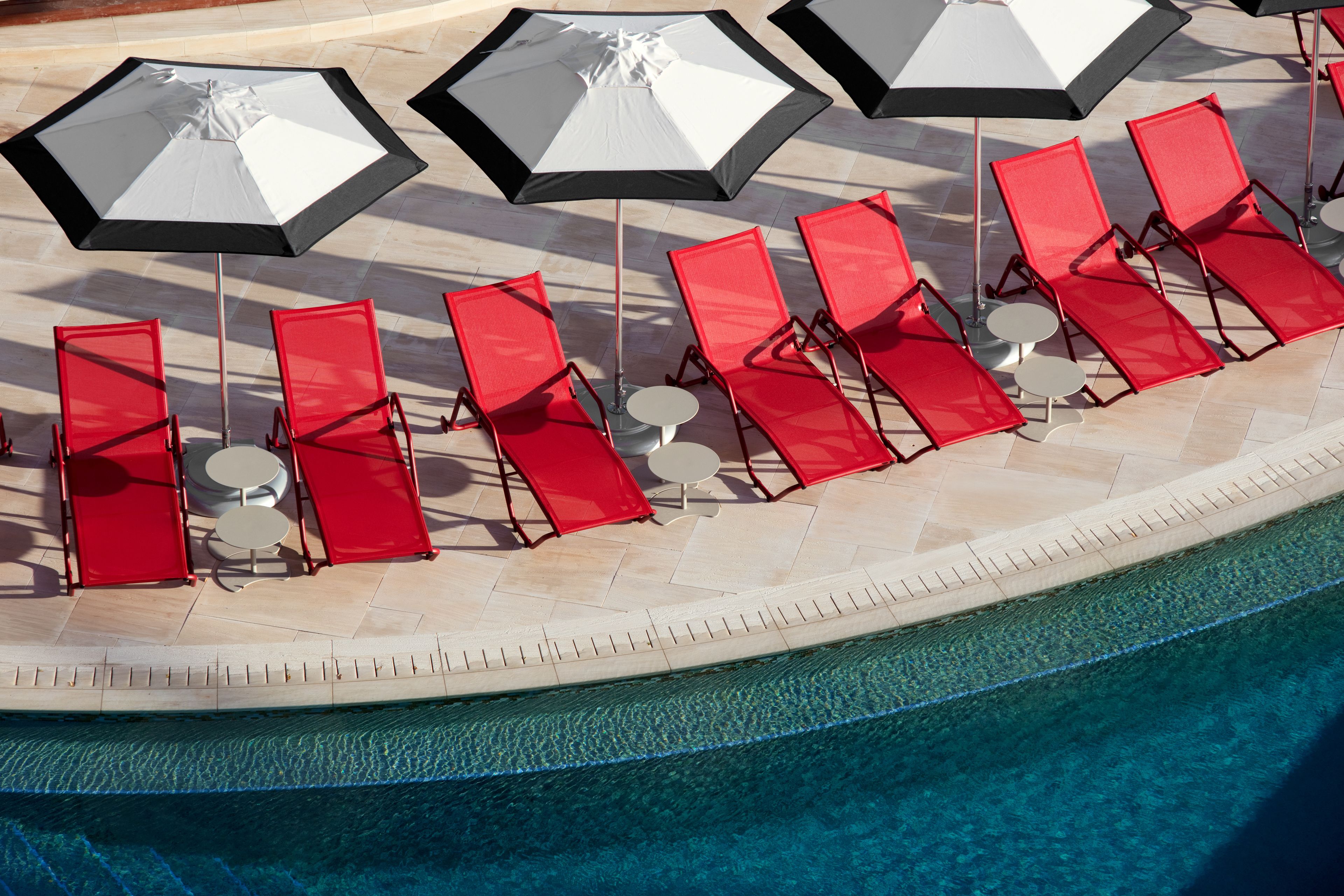 A poolside area with red lounge chairs and large umbrellas, a tiled deck, and a clear blue pool.