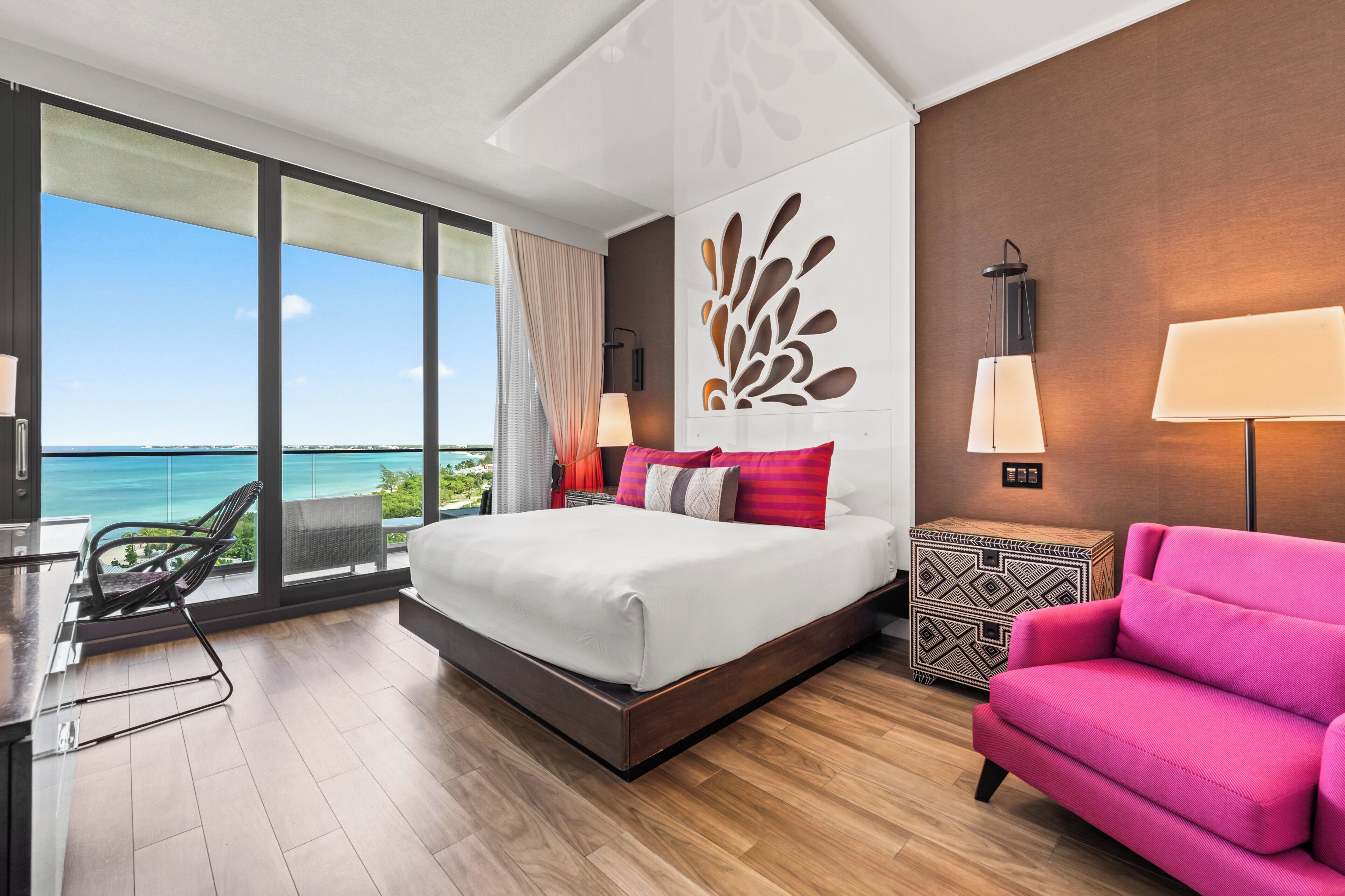 A modern hotel room with a large bed, a view of the sea, a balcony with a chair, and a pink armchair.