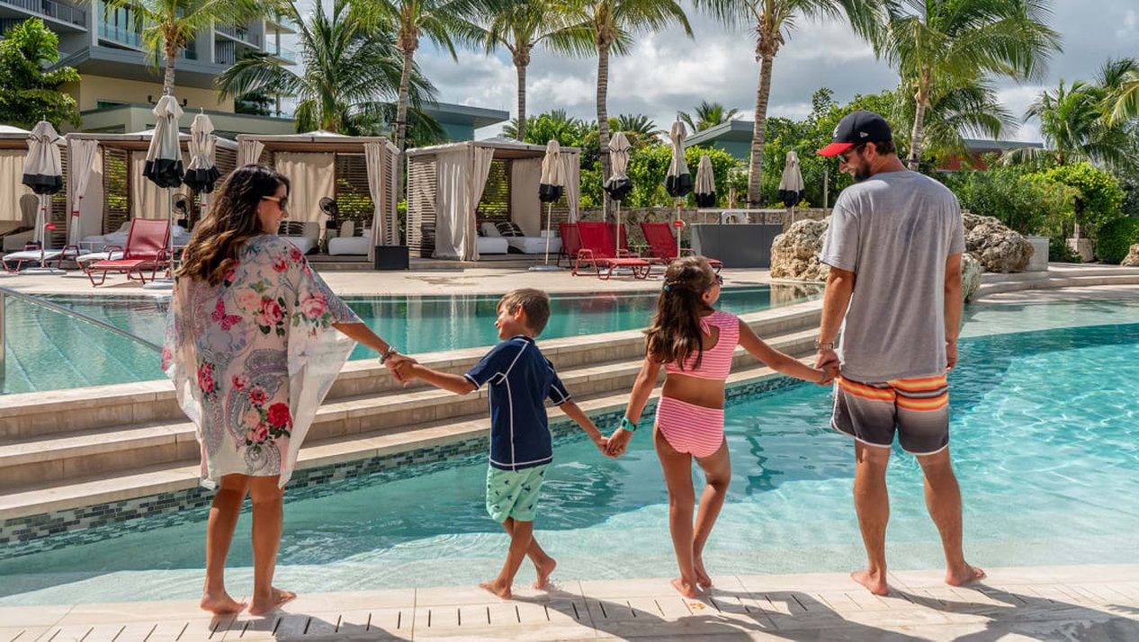 Una familia tomándose de las manos junto a una piscina, con sillones de descanso y sombrillas al fondo.