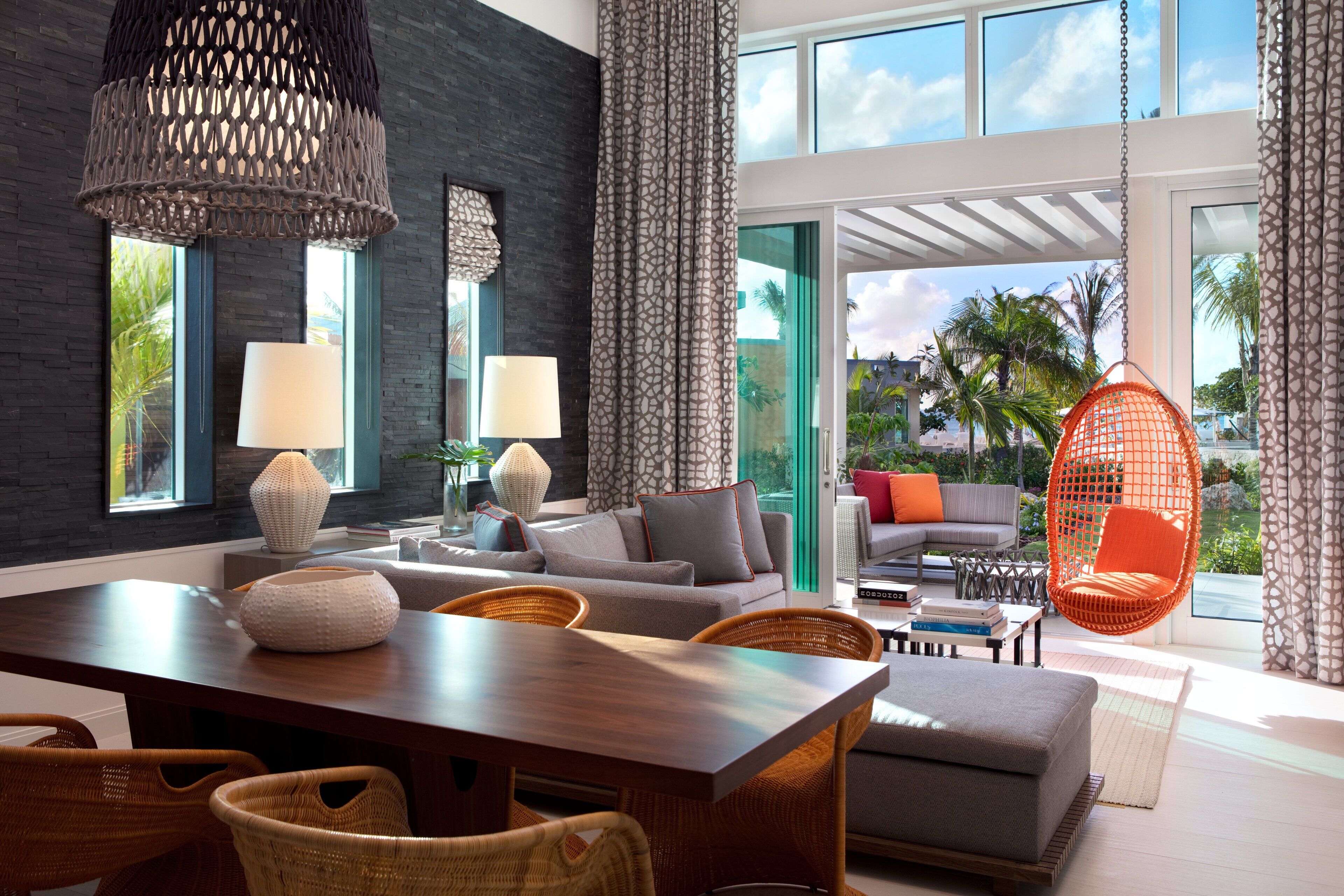 A modern living room with a wooden dining table, wicker chairs, a grey sofa, and a hanging orange chair.