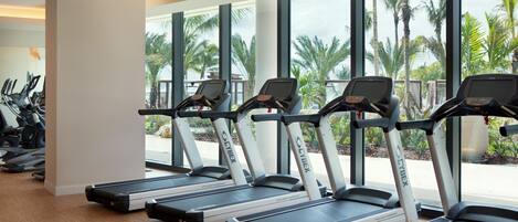 A gym with treadmills aligned against a wall with large windows overlooking palm trees.