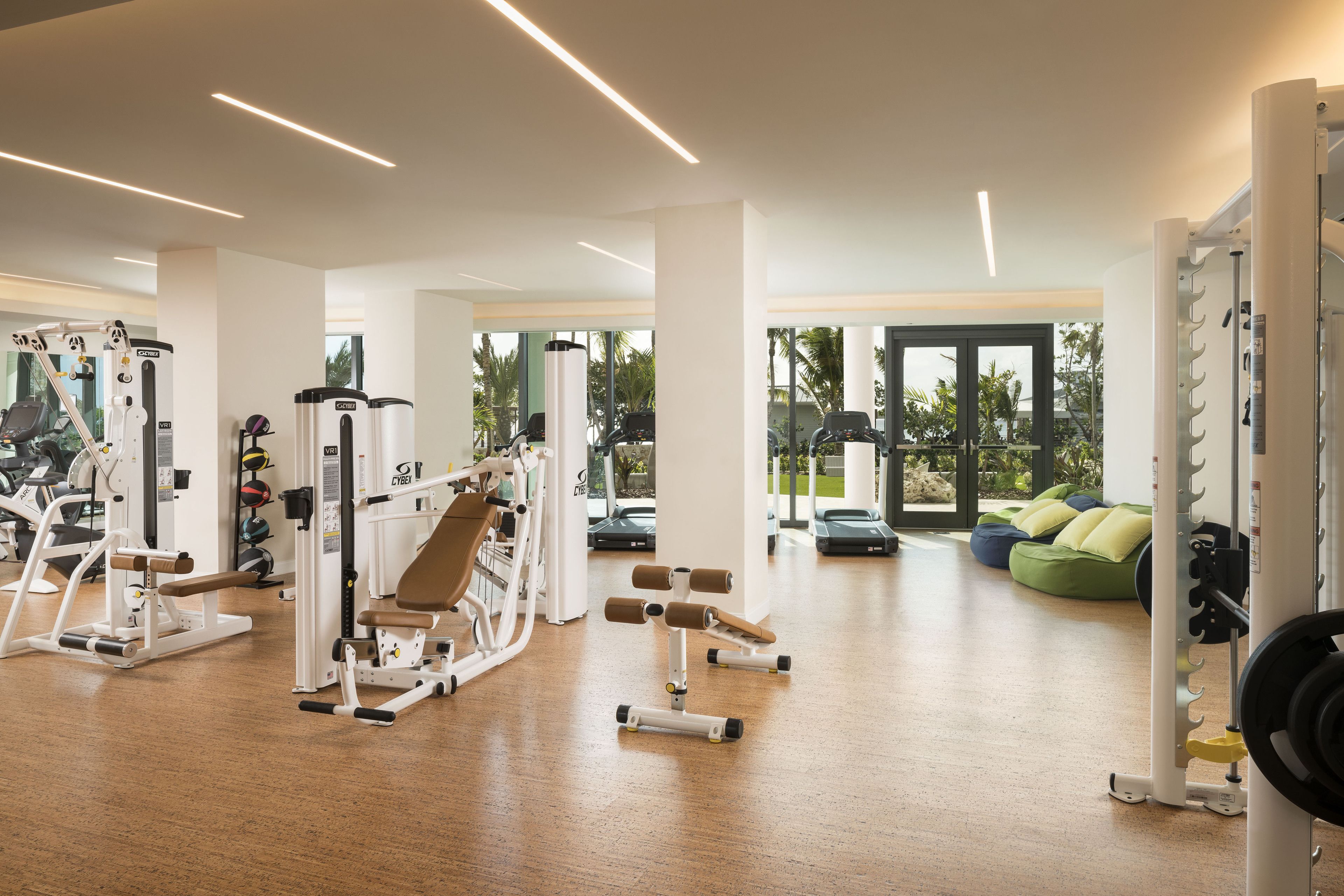 Een ruime fitnessruimte met diverse trainingsapparaten, grote ramen en een groene relaxfauteuil.