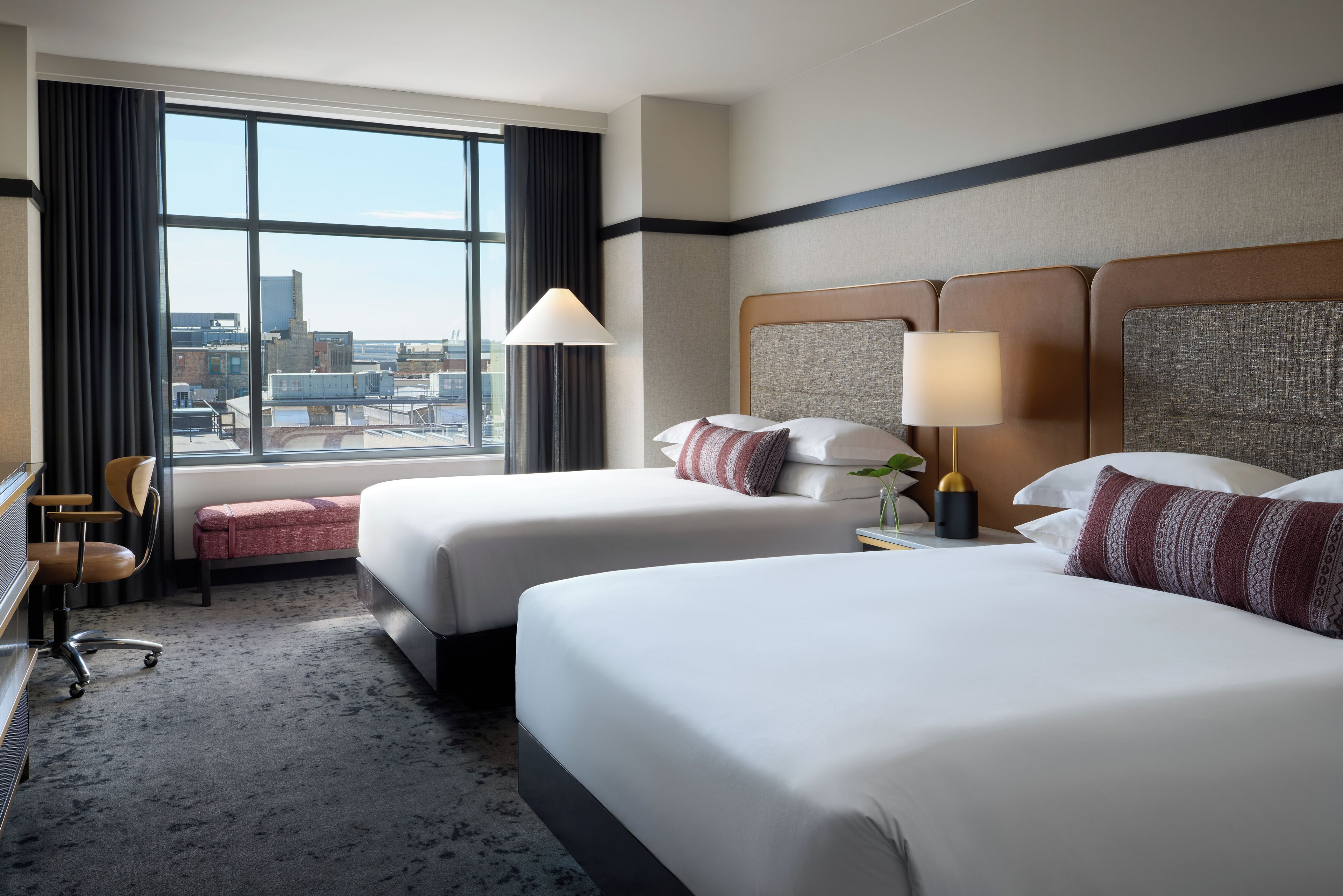 A hotel room with two beds, a desk, a chair, a window with curtains, and a view of the cityscape.