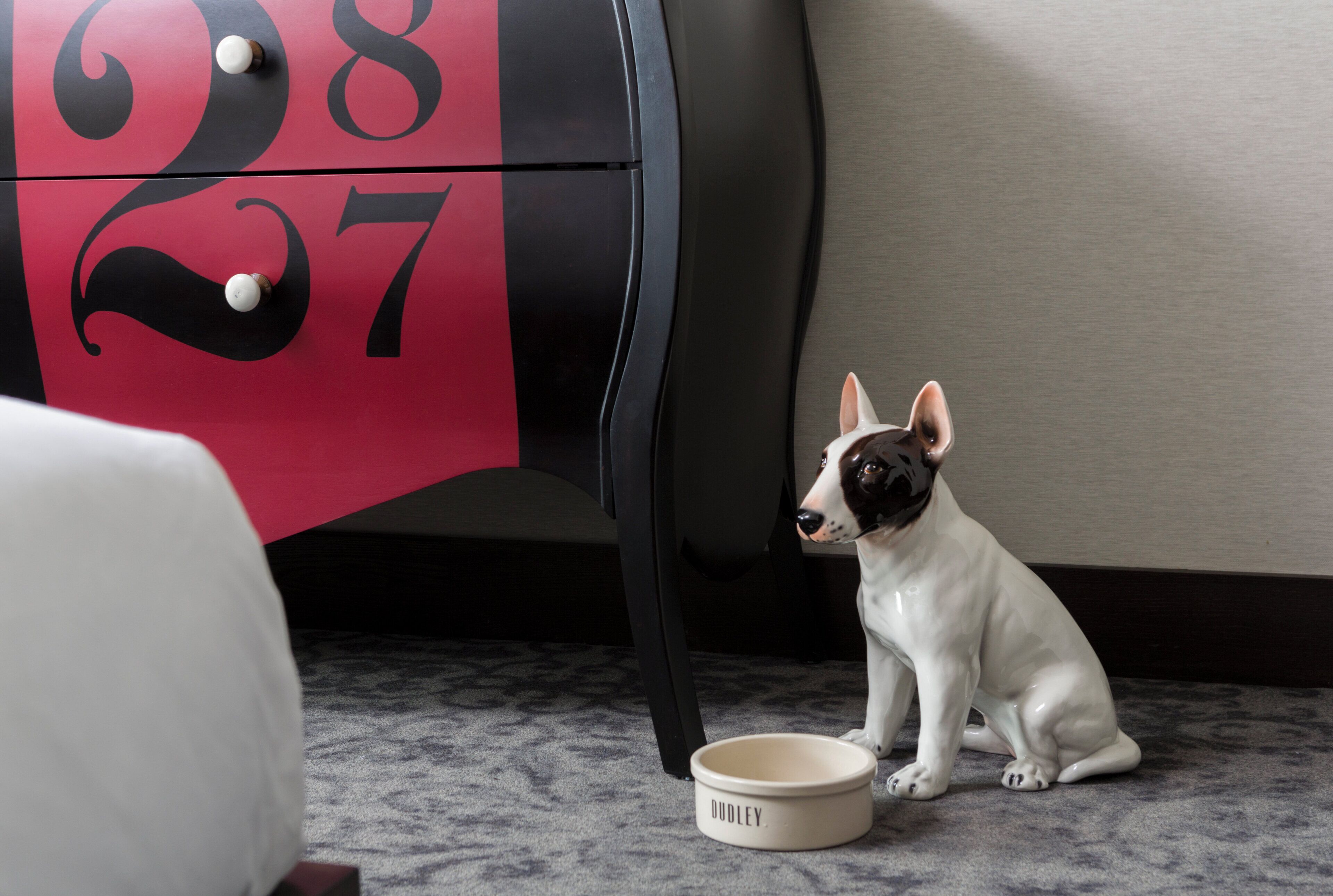 A small white dog sitting next to a bowl on a carpeted floor, with a red and black cabinet in the background.