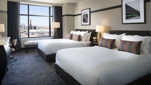 A hotel room with two beds, a desk, and a large window offering a city view. - Kimpton Journeyman Hotel by IHG (Milwaukee)