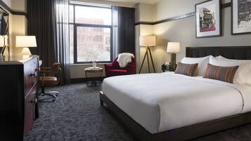 A hotel room with a large bed, two bedside tables with lamps, a desk with a chair, a red armchair, and a view of a cityscape through the window.