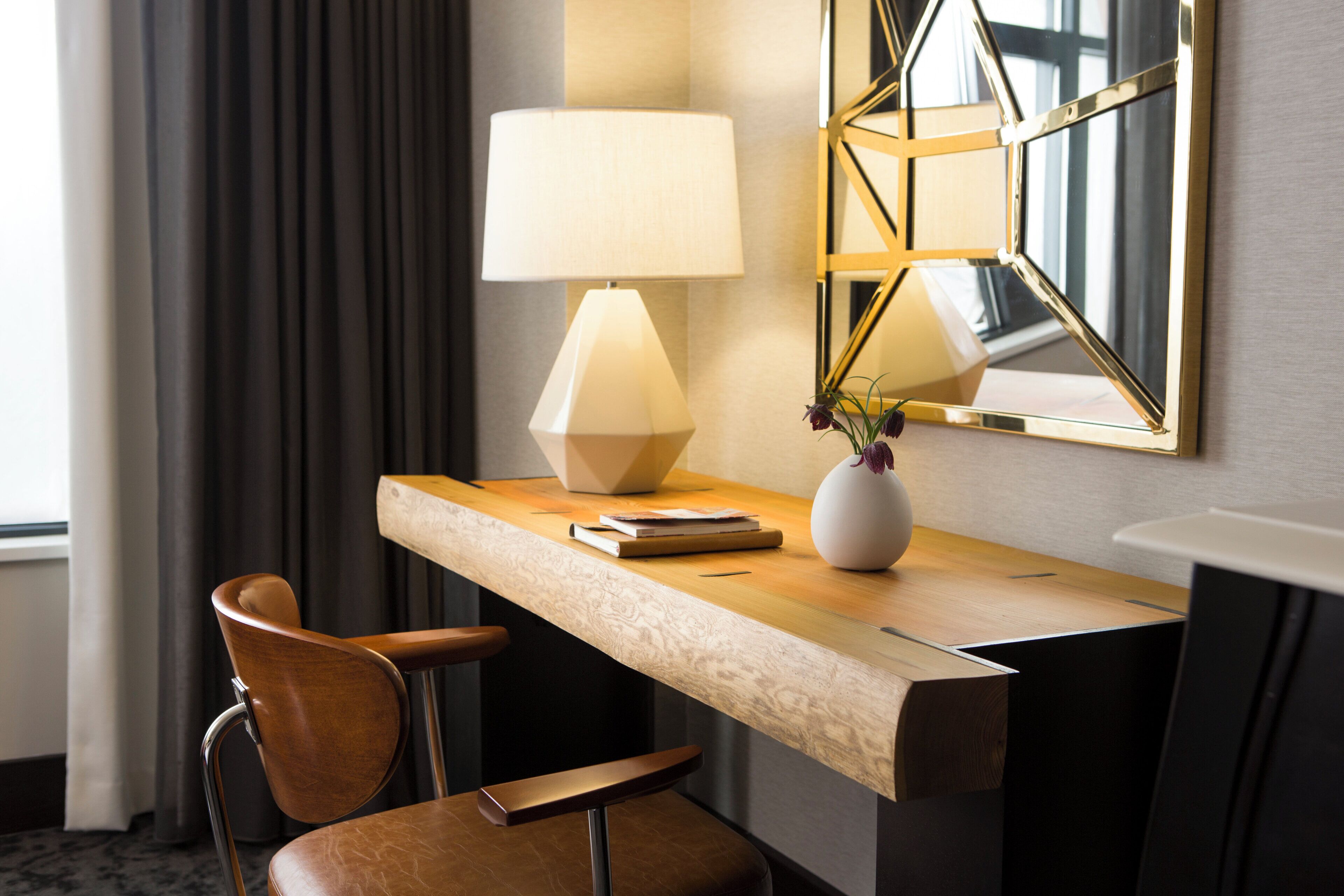 Habitación de hotel con escritorio de madera, una silla, una lámpara, un jarrón con flores y un espejo en la pared.