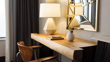 Habitación de hotel con escritorio de madera, una silla, una lámpara, un jarrón con flores y un espejo en la pared.