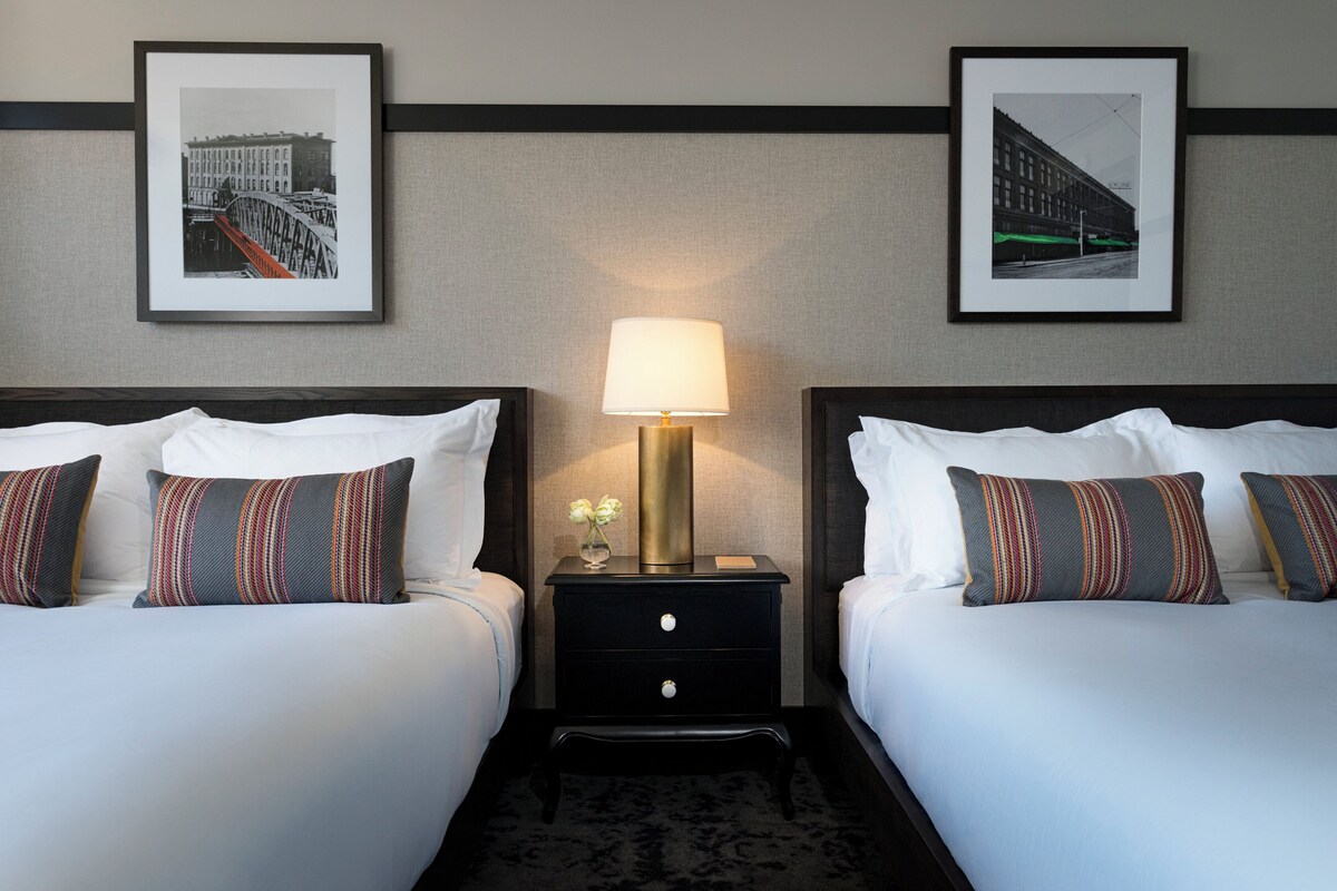 A hotel room with two beds, a nightstand with a lamp, and two framed pictures on the wall.