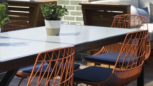 An outdoor dining area with a table and orange wireframe chairs, a potted plant, and a brick wall in the background. - Kimpton Journeyman Hotel by IHG (Milwaukee)