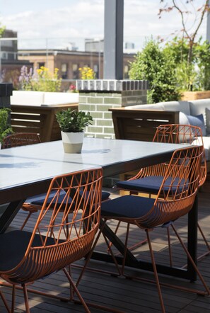 An outdoor dining area with a table and orange wireframe chairs, a potted plant, and a brick wall in the background. - Kimpton Journeyman Hotel by IHG (Milwaukee)