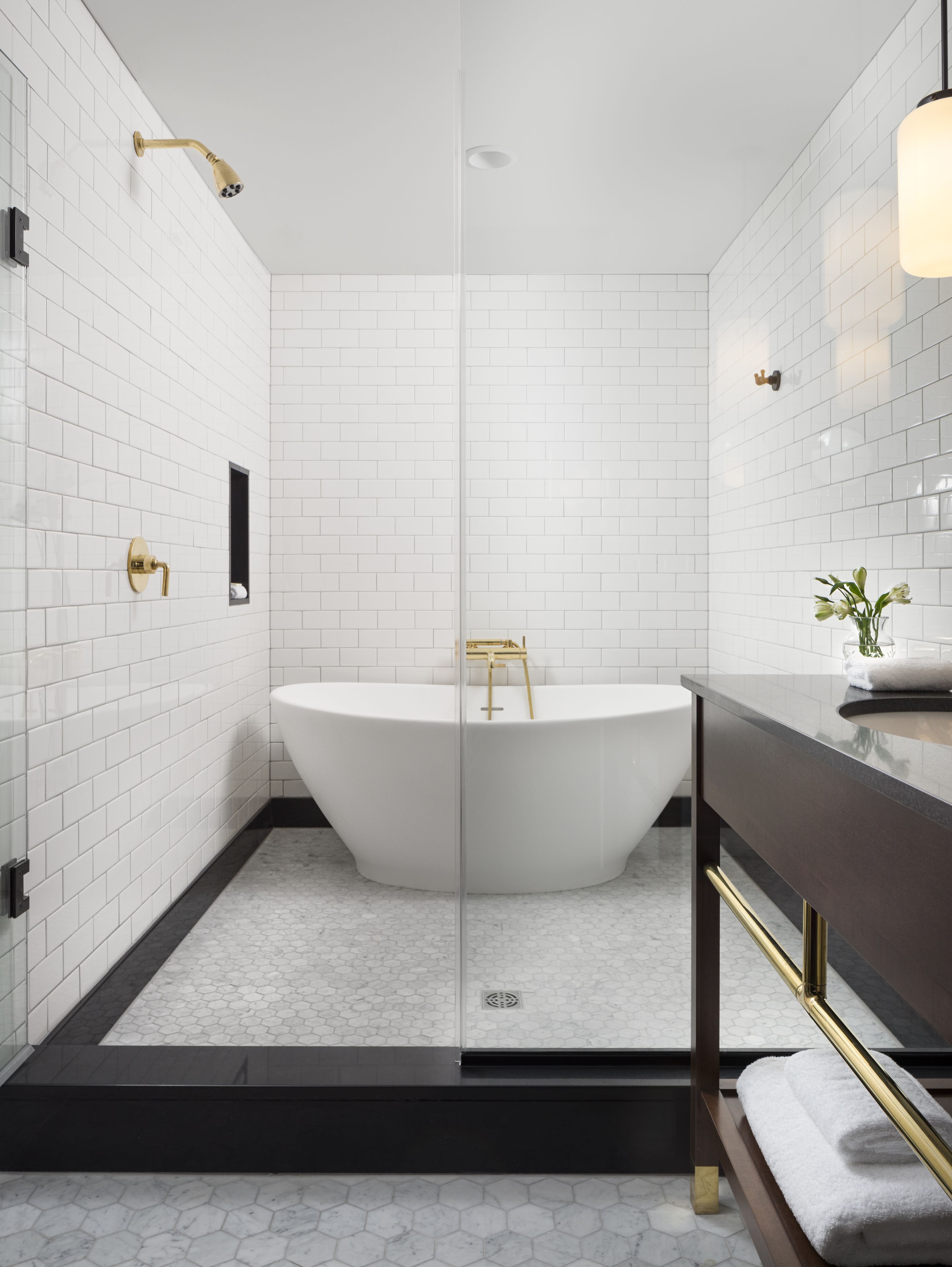 Un baño moderno con azulejos blancos tipo metro, una bañera independiente, un box de ducha con mampara de vidrio y un mueble de tocador de madera oscura con detalles dorados.