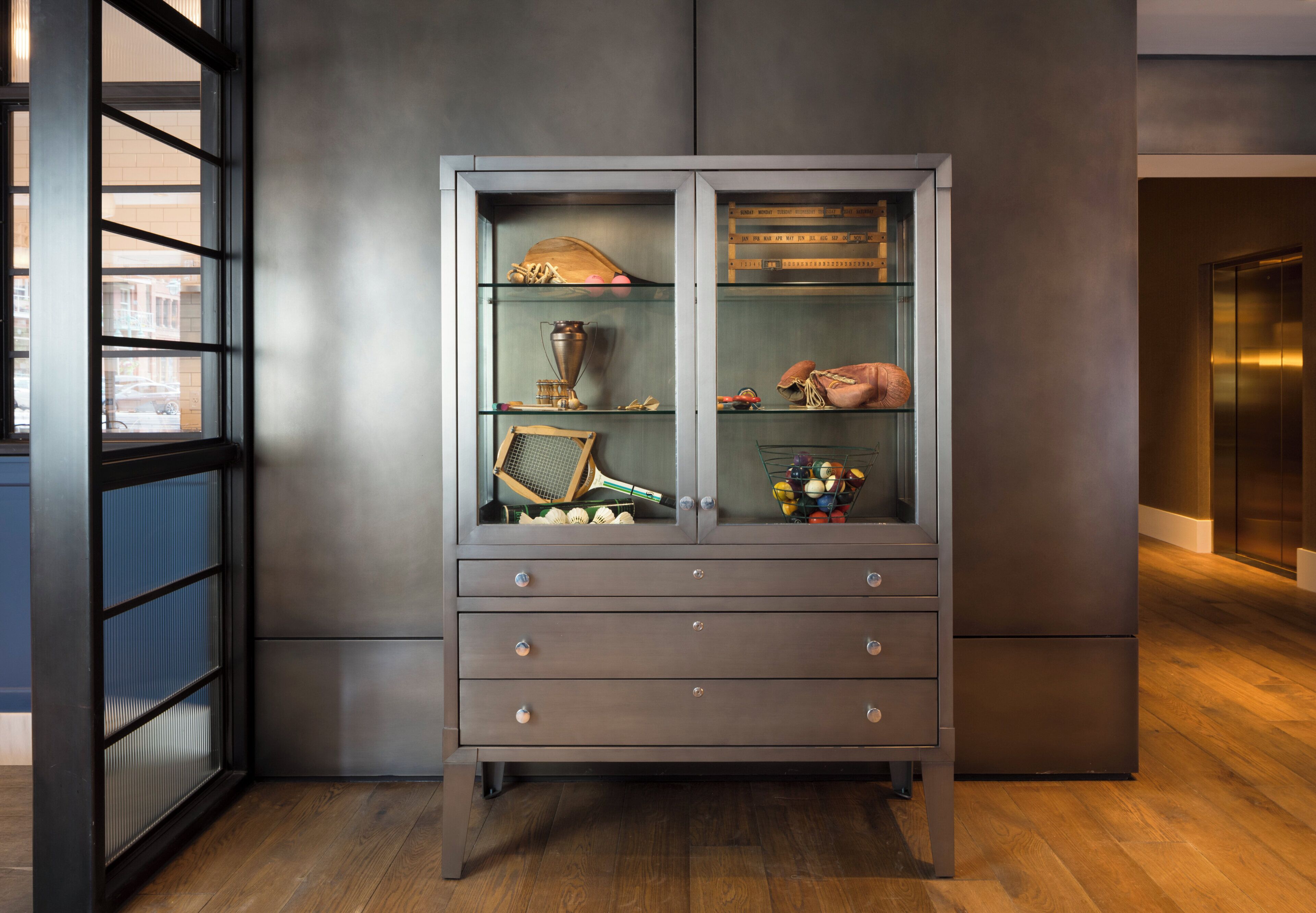 A dark wooden cabinet with glass doors, containing various items like a trophy, a tennis racket, and a pair of shoes.