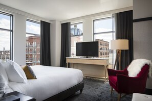 A hotel room with a large bed, a desk with a TV, a red chair, and a view of the city through large windows. - Kimpton Journeyman Hotel by IHG (Milwaukee)