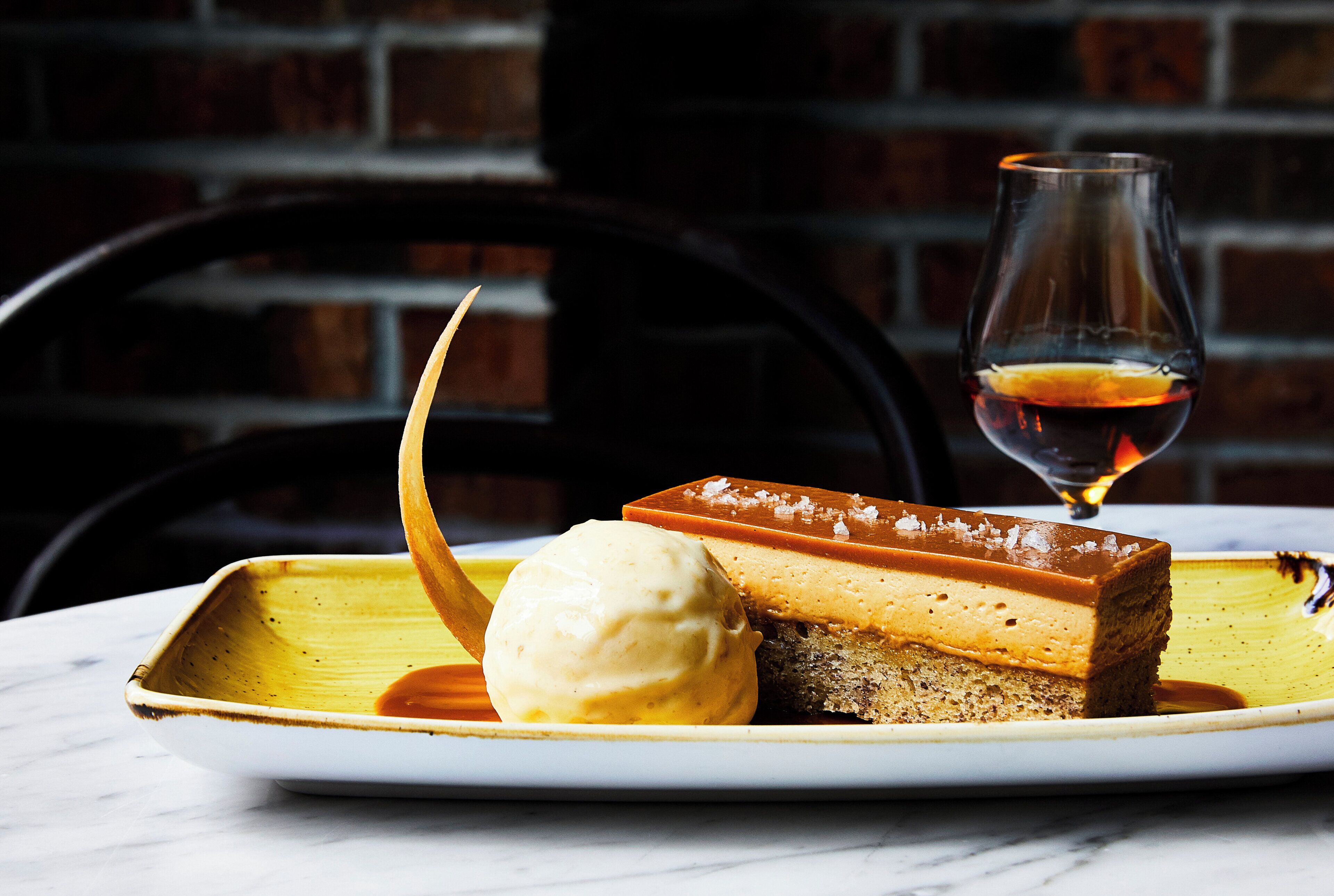 Un plato de postre con una porción de pastel, una bola de helado y un vaso de un líquido oscuro, posiblemente un vino dulce, sobre un plato blanco con borde amarillo.